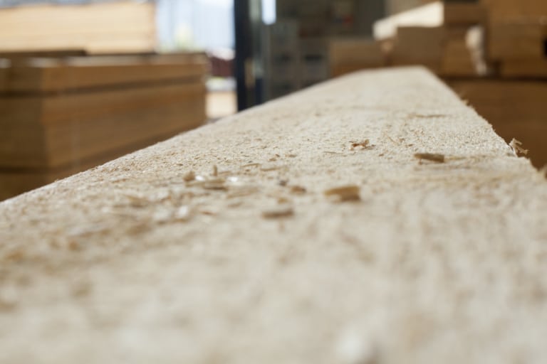 Eine Nahaufnahme eines langen Holzbretts, das mit Holzspänen bedeckt ist. Die Oberfläche des Brettes zeigt eine raue Textur und die Maserung des Holzes. Im Hintergrund sind unscharfe Holzstapel sichtbar, die auf eine Produktionsumgebung hinweisen. Das Licht ist hell und betont die natürlichen Farben des Holzes.