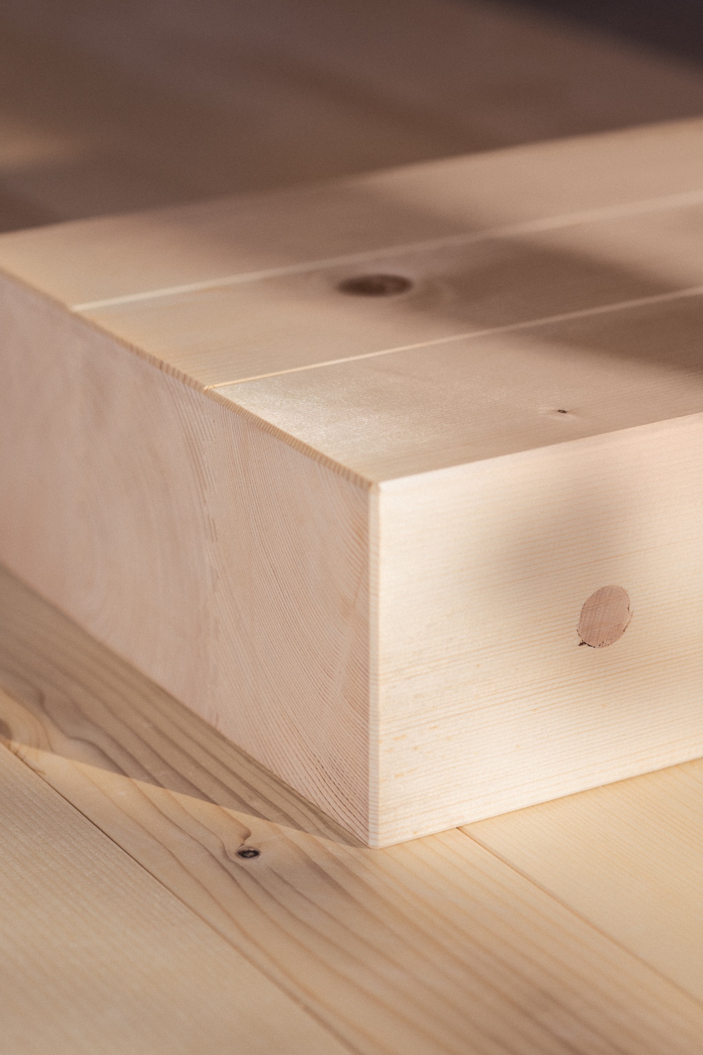 Eine Vollholzdecke von holzius, die aus mehreren massiven Holzplatten besteht. Die Oberfläche ist glatt und zeigt eine helle, natürliche Holzmaserung mit kleinen Knoten. Ein Kantenbereich ist deutlich sichtbar, während der Hintergrund aus weiteren Holzoberflächen besteht, die die hochwertige Verarbeitung des Materials betonen.