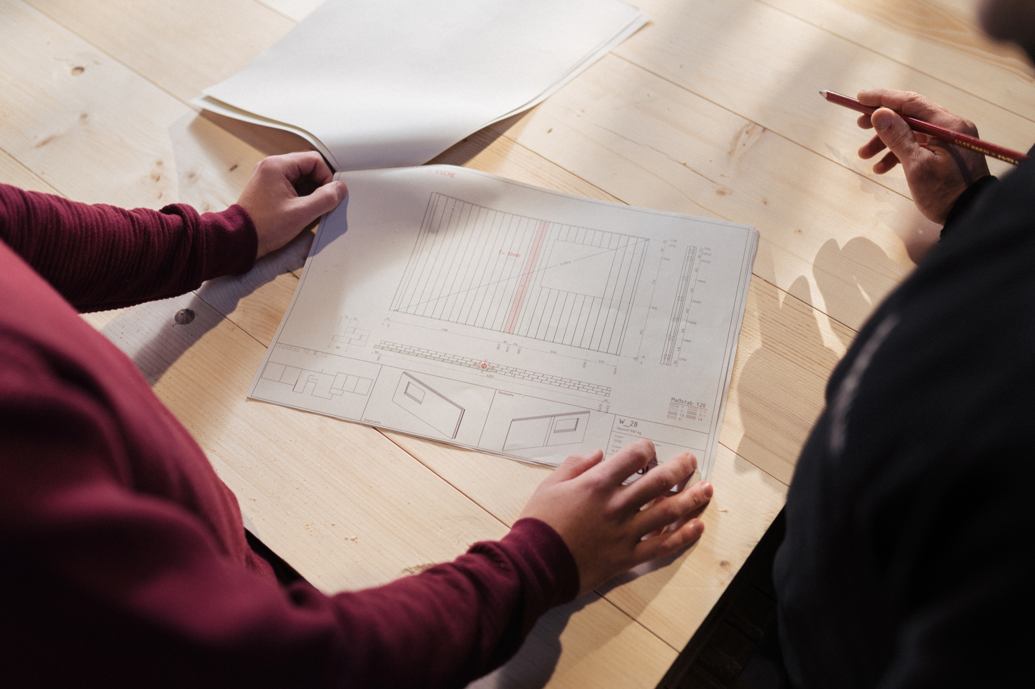 Zwei Personen sitzen an einem Holztisch und besprechen Produktionspläne für Wände. Vor ihnen liegt ein detaillierter Plan mit technischen Zeichnungen und Notizen. Eine Person hält einen Bleistift und zeigt auf einen spezifischen Bereich des Plans, während die andere Hand das Dokument festhält. Der Tisch hat eine helle Holzoberfläche und der Raum ist gut beleuchtet.