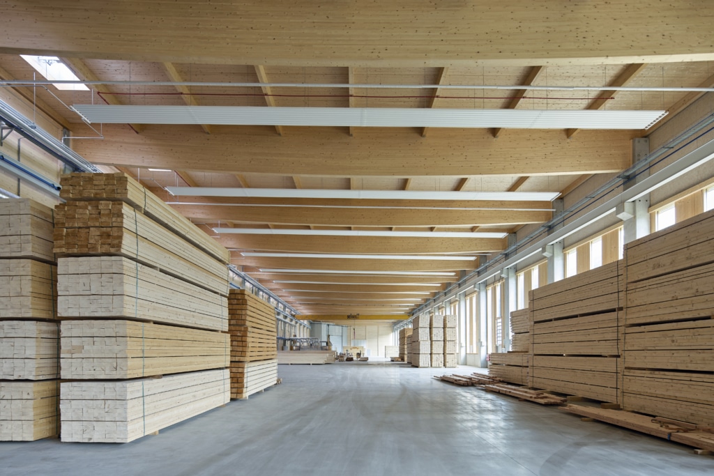 Eine geräumige Lagerhalle mit gestapelten Holzbrettern auf beiden Seiten. Die Decke besteht aus großen Holzbalken, und der Boden ist aus Beton. Rechts befinden sich große Fenster, die viel Tageslicht hereinlassen. Die Halle wirkt sauber und gut organisiert.