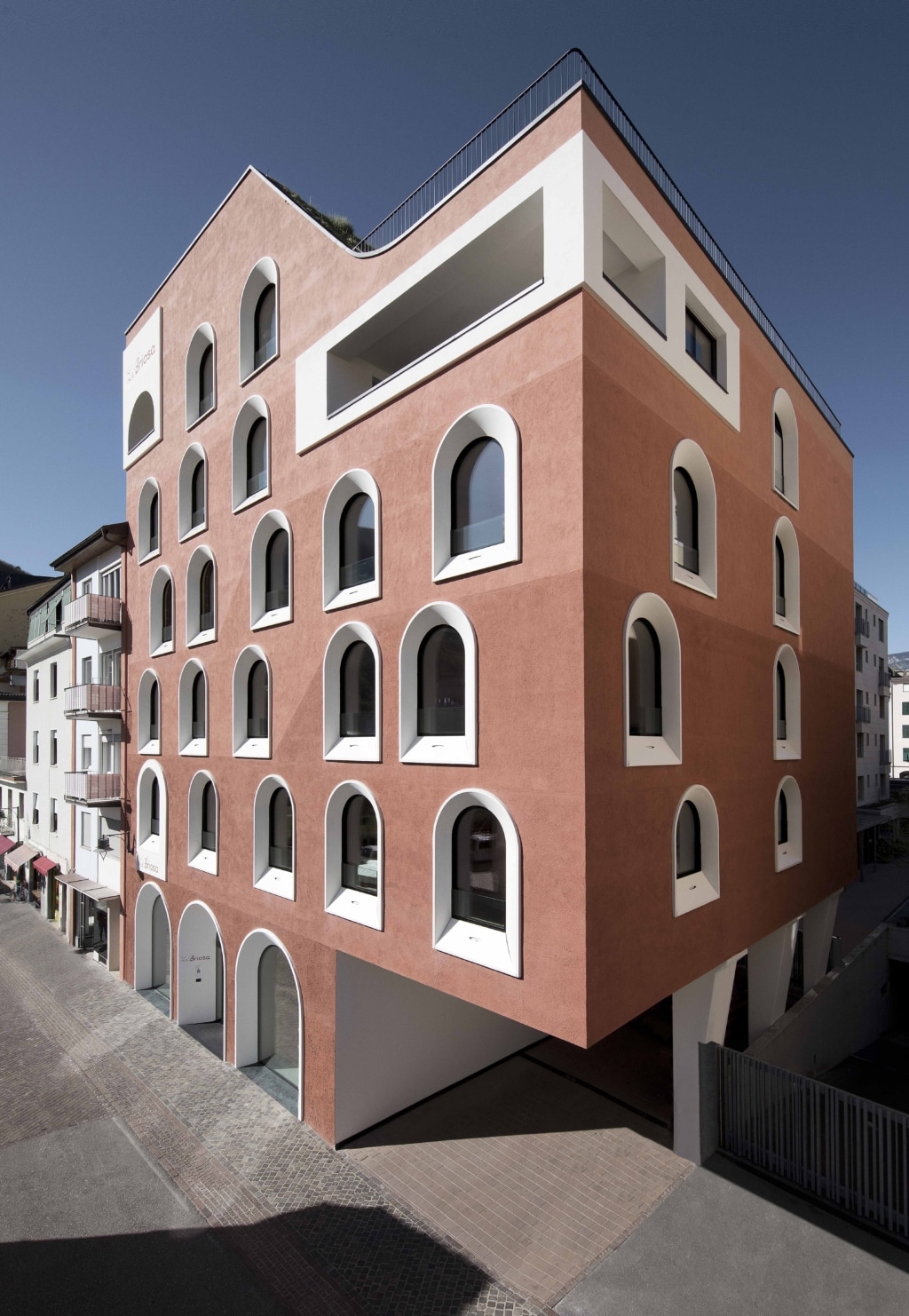 Außenansicht eines markanten, modern gestalteten Gebäudes mit terrakottafarbener Fassade und klarer, kubischer Form. Die Fassade ist regelmäßig mit hohen, schmalen Rundbogenfenstern gegliedert, die von weißen, plastisch hervorgehobenen Rahmen eingefasst sind und einen starken Kontrast zur warmen Farbfläche bilden. Das Gebäude steht an einer Straßenecke und ruht teilweise auf schlanken Stützen, wodurch im Erdgeschoss ein offener Bereich entsteht. In den oberen Etagen sind zusätzliche Einschnitte und eine umlaufende Brüstung zu erkennen. Links schließen sich angrenzende Wohnhäuser mit Balkonen an. Der Straßenbereich ist gepflastert, und der klare blaue Himmel unterstreicht die klare, reduzierte Architektur und die prägnante Farbwirkung des Baukörpers.