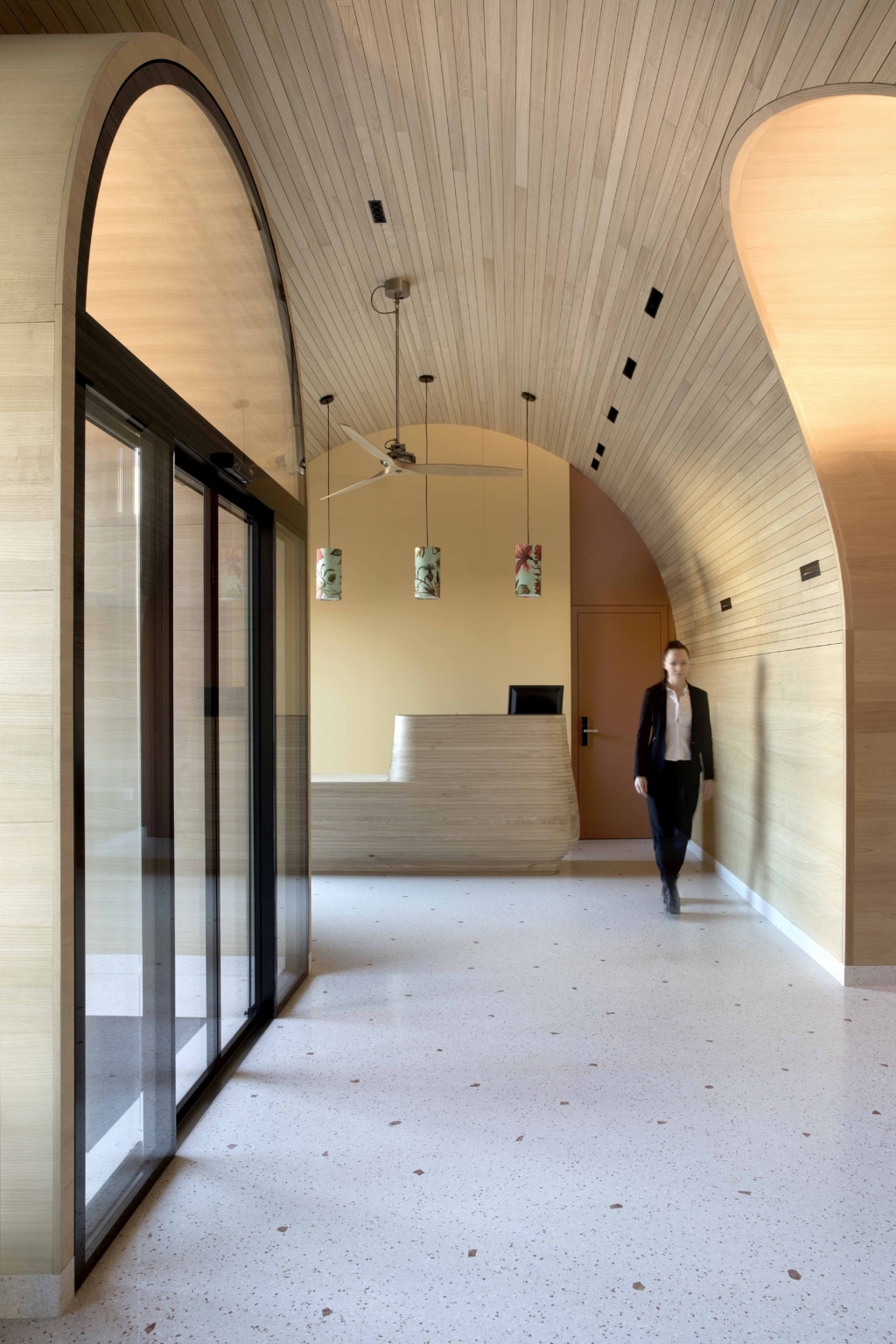 Moderner Empfangsbereich mit fließender architektonischer Gestaltung und warmen Naturmaterialien: Ein langer, heller Flur mit gesprenkeltem Boden führt zu einer organisch geformten Rezeption aus hellem Holz, die vor einer in warmem Beige gehaltenen Wand positioniert ist. Die Decke ist vollständig mit feinlamelliertem Holz verkleidet und wölbt sich in weichen, geschwungenen Formen über den Raum. Mehrere Pendelleuchten mit gemusterten Schirmen hängen in einer Linie über dem Empfangstresen, ergänzt durch einen großen, langsam rotierenden Deckenventilator. Auf der linken Seite erstreckt sich eine durchgehende Glasfront mit großen Schiebetüren, die viel Tageslicht in den Raum lässt und Ausblicke nach außen ermöglicht. Rechts führt ein Durchgang mit ebenfalls geschwungenem Holzrahmen weiter. Im Vordergrund bewegt sich eine Person durch den Raum, was dem ansonsten ruhigen, minimalistischen Setting eine lebendige Note verleiht. Die Kombination aus Holz, weichen Rundungen, warmen Farbtönen und klaren Linien schafft eine einladende, moderne und hochwertige Atmosphäre.
