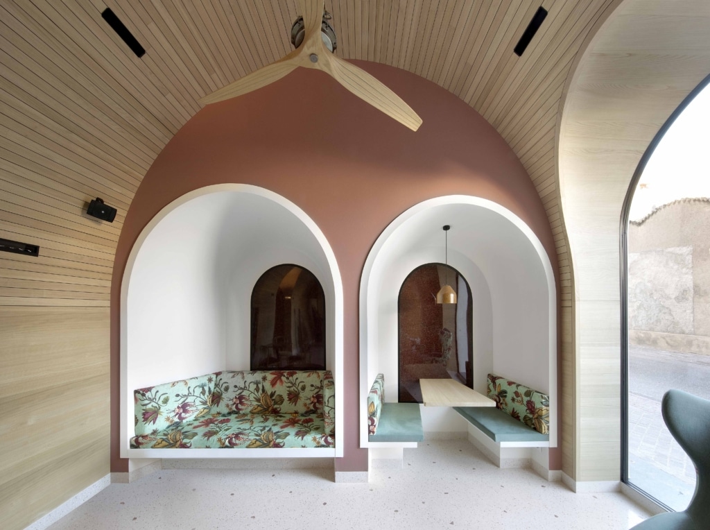 Architektonisch gestalteter Lounge- oder Aufenthaltsbereich mit klaren Rundformen und warmen Naturmaterialien: Zwei großzügige Sitznischen mit halbkreisförmigen Öffnungen sind symmetrisch in eine terrakottafarbene Wand eingelassen. Die linke Nische ist mit einer durchgehenden Polsterbank in floral gemustertem Stoff ausgestattet und wird durch einen kleinen runden Holztisch ergänzt. In der rechten Nische befinden sich zwei gegenüberliegende Sitzbänke mit schlichtem, grünem Bezug und ein schmaler Tisch, darüber hängt eine dezente Pendelleuchte. Die Decke ist mit fein strukturiertem Holz verkleidet und wird von einem großen Deckenventilator mit geschwungenen Holzflügeln dominiert. Rechts sorgt eine große, gebogene Fensterfläche für viel Tageslicht und gibt den Blick nach draußen frei. Der helle, gesprenkelte Boden und die harmonische Kombination aus Holz, warmen Farbtönen und weichen Formen erzeugen eine ruhige, einladende und hochwertig gestaltete Atmosphäre.
