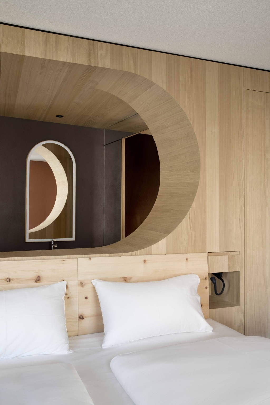 Detailansicht eines modern gestalteten Hotelzimmers mit Fokus auf die architektonische Innenraumgestaltung: Im Vordergrund ein Doppelbett mit weißer Bettwäsche und einem schlichten Kopfteil aus hellem Holz mit sichtbarer Maserung. Dahinter erstreckt sich eine großflächige Holzverkleidung, die in eine markante, halbkreisförmige Öffnung übergeht und den Blick in einen angrenzenden Bereich lenkt. In dieser Öffnung ist ein Spiegel mit abgerundeter Oberkante zu sehen, der warme Terrakottafarben und weitere architektonische Rundformen reflektiert. Rechts befindet sich eine integrierte Nische mit kleiner Leuchte, links eine glatte, dunklere Wandfläche als Kontrast. Die Kombination aus hellen Holztönen, weichen Rundungen und warmen Farben schafft eine ruhige, elegante und hochwertig gestaltete Atmosphäre.