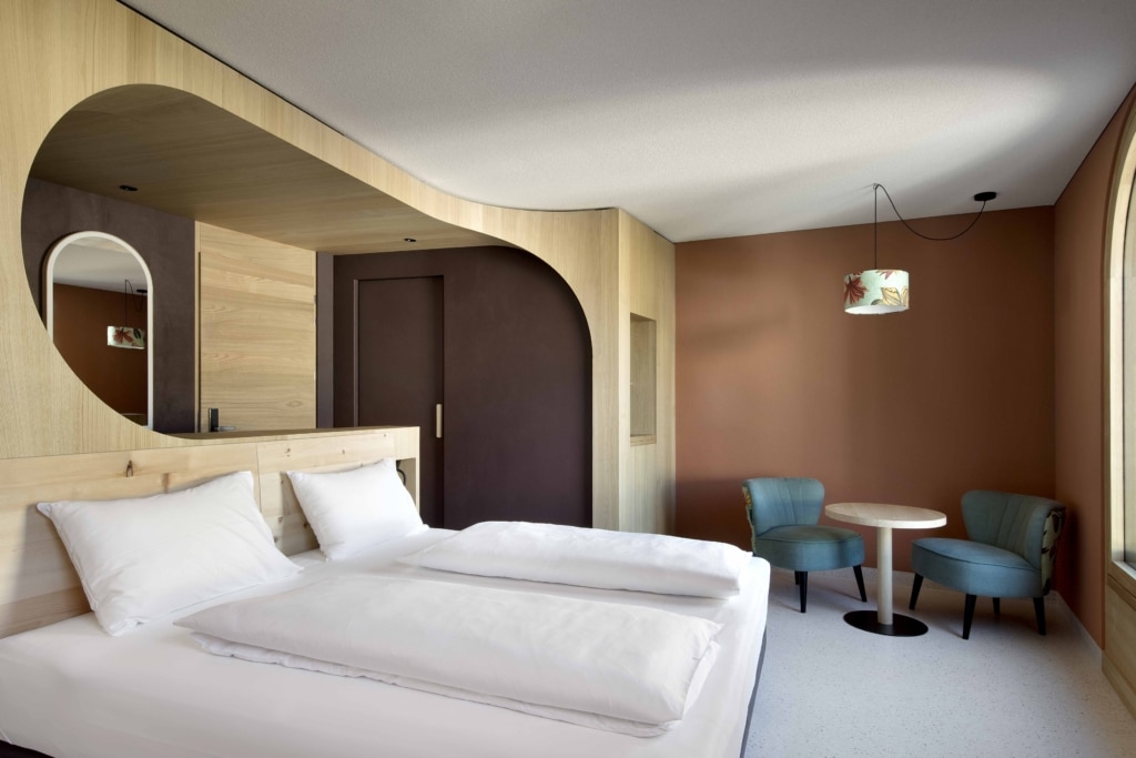 Modernes Hotelzimmer mit klarer, reduzierter Gestaltung und warmen Naturmaterialien: Im Vordergrund ein großzügiges Doppelbett mit weißer Bettwäsche und hellen Holzelementen am Kopfteil. Dahinter zieht sich eine fließend geschwungene Holzverkleidung über Wand und Decke und bildet weiche architektonische Übergänge. Eine dunklere Wandfläche und ein integrierter Durchgang schaffen Tiefe im Raum. Auf der rechten Seite befindet sich eine gemütliche Sitzecke mit zwei gepolsterten Sesseln in gedämpftem Grün und einem runden Holztisch vor einer terrakottafarbenen Wand. Eine hängende Leuchte mit dekorativem Stoffschirm setzt einen farblichen Akzent. Große Fenster lassen viel Tageslicht in den Raum und unterstreichen die ruhige, elegante Atmosphäre.