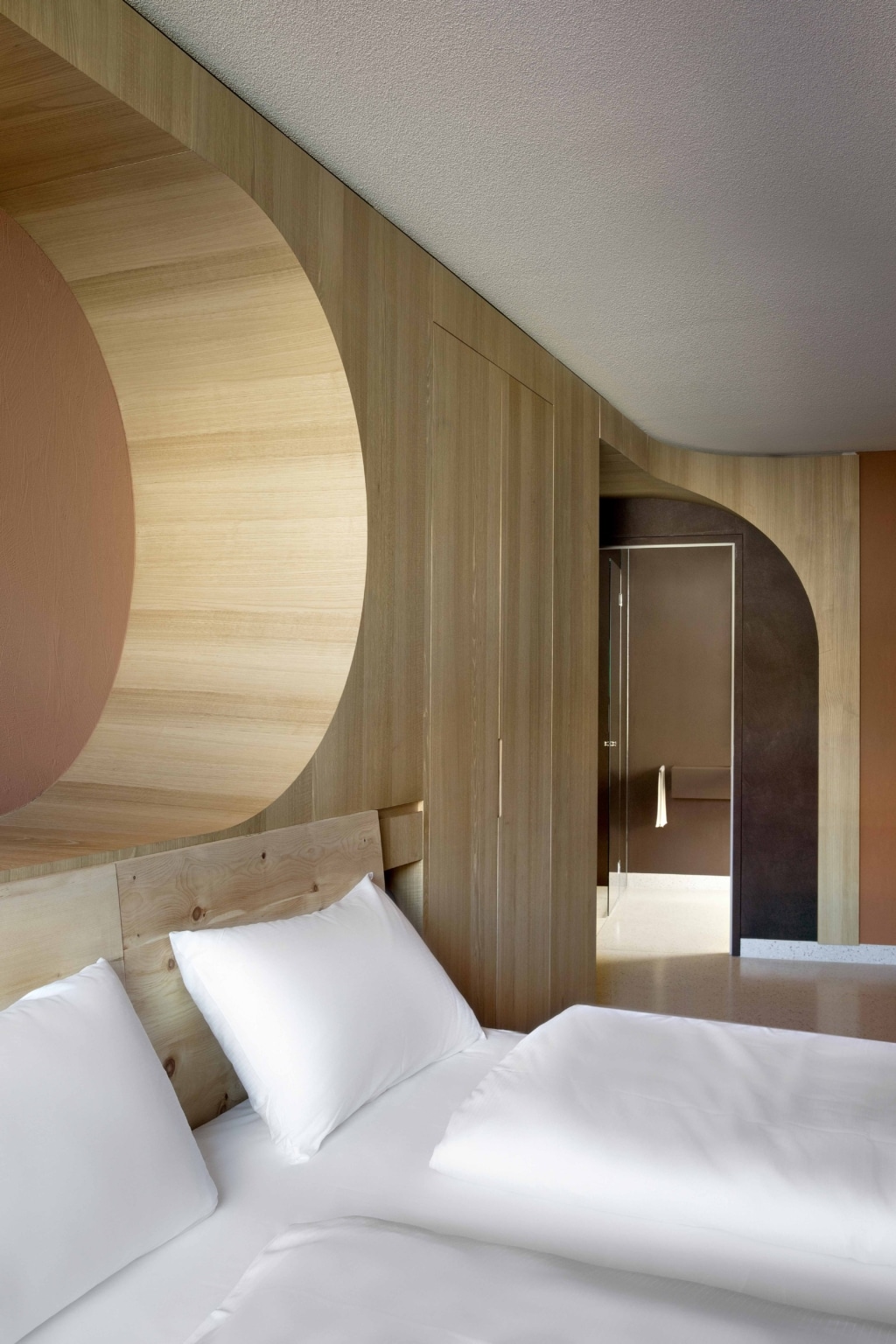 Detailaufnahme eines modernen Hotelzimmers mit warmem, minimalistischem Design: Im Vordergrund ein Bett mit weißer Bettwäsche und Kissen, dahinter eine helle Holzvertäfelung, die sich über die Wand und in fließenden Linien bis zur Decke zieht. In die Holzverkleidung ist ein großes, rundes architektonisches Element integriert, das eine weiche, organische Form bildet. Rechts führt eine bogenförmige Öffnung in einen angrenzenden Raum mit Tür und indirekter Beleuchtung. Die Farbpalette besteht aus natürlichen Holztönen, warmen Erdfarben und gedeckten Rosé- und Braunnuancen, die eine ruhige und elegante Atmosphäre schaffen.