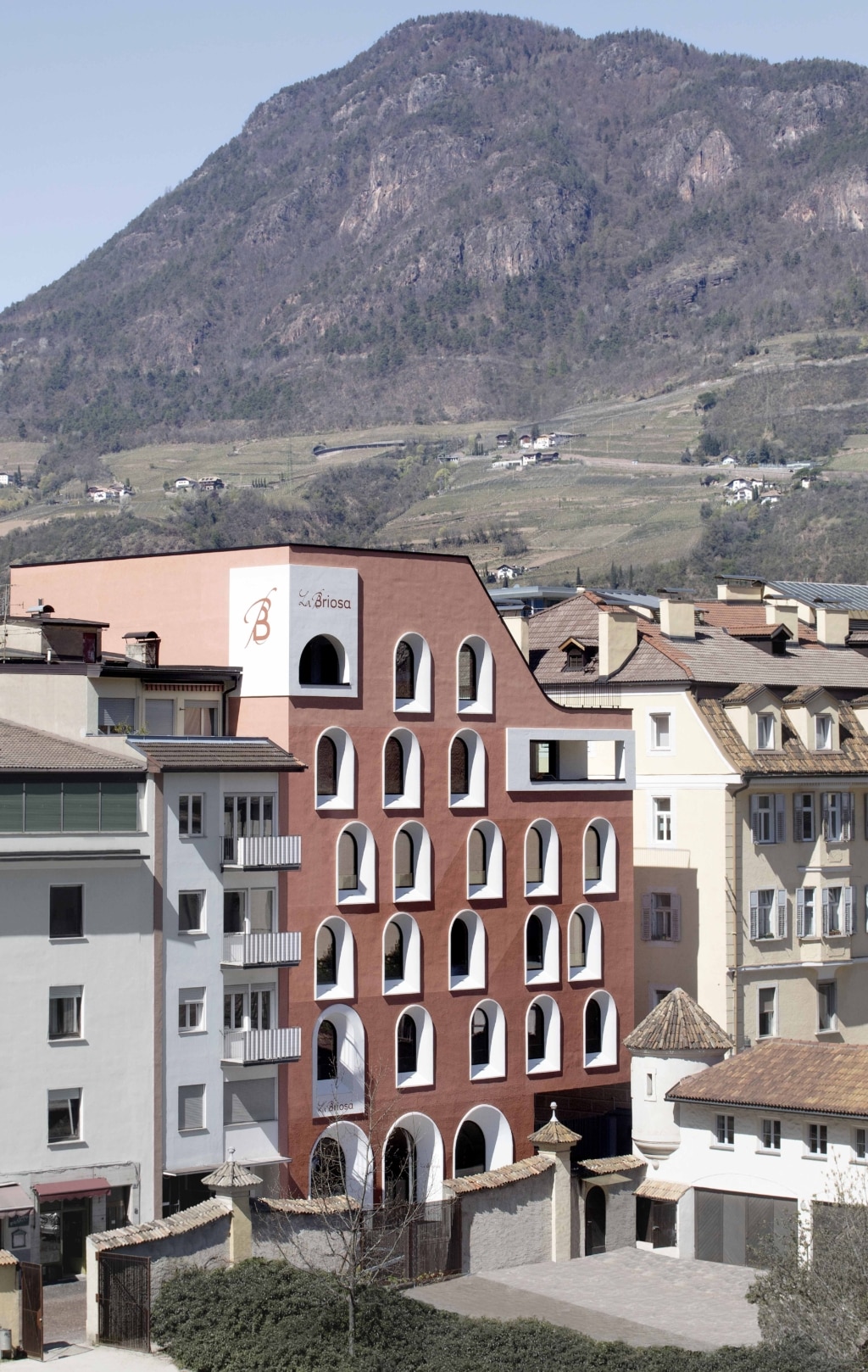 Mehrstöckiges, modernes Gebäude in terrakottafarbener Fassade mit zahlreichen symmetrisch angeordneten, bogenförmigen Fenstern und weiß abgesetzten Rahmen. Das Haus trägt den Schriftzug ‚La Briosa‘ und befindet sich eingebettet zwischen hellen Wohnhäusern mit Ziegeldächern. Im Vordergrund ein ummauerter Hofbereich mit kleinem Türmchen, im Hintergrund erstreckt sich ein steiler, teils bewaldeter Berghang mit Weinbergen und vereinzelten Höfen unter klarem Himmel in alpiner Umgebung.