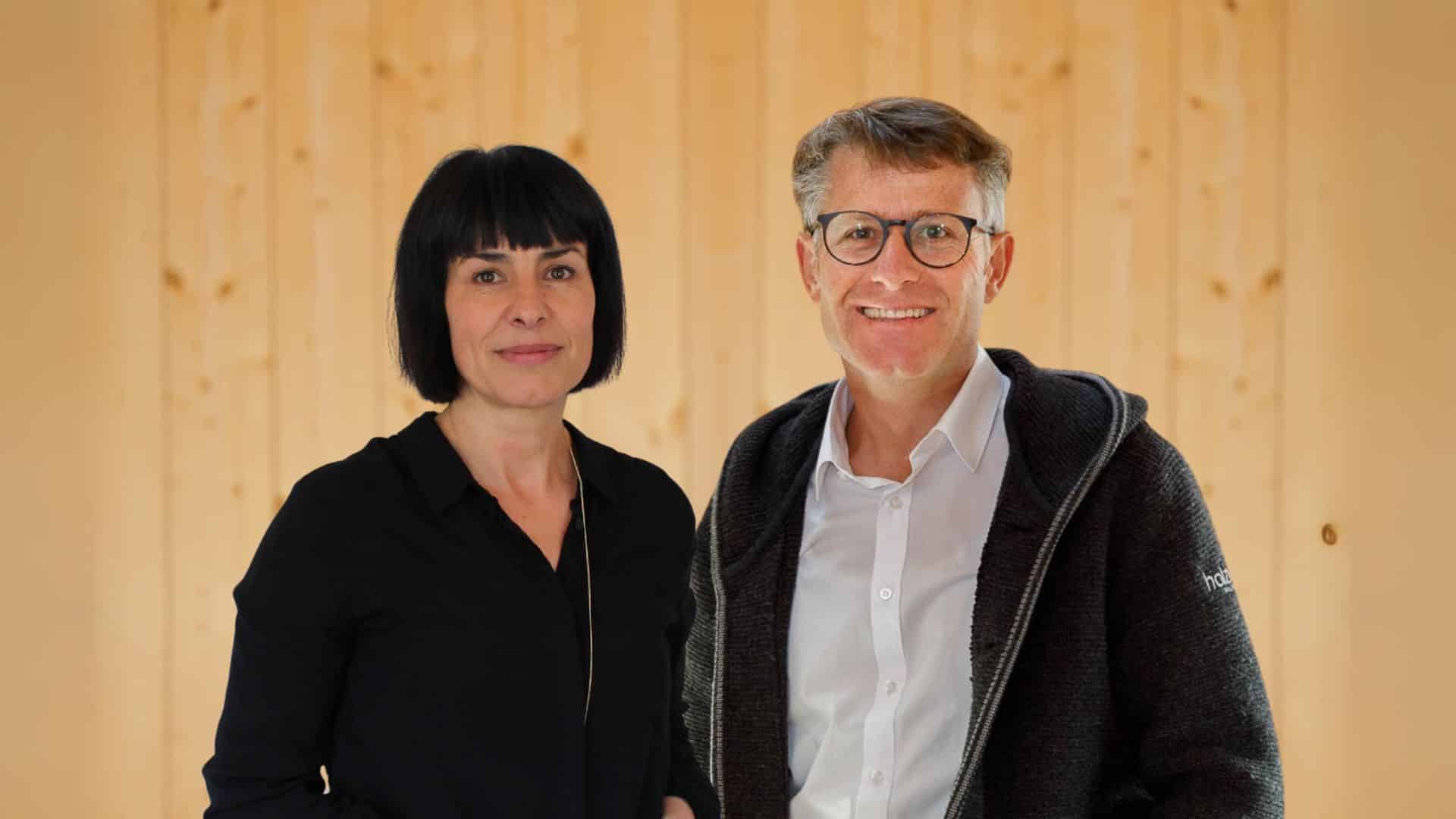 Au dem Bild sieht man zwei Personen, die vor einem Holzhintergrund stehen. Links steht Maria Unterfrauner, holzius Marketingleiterin. Sie hat dunkle halblange Haare und trägt eine schwarze Bluse. Rechts neben ihr steht holzius-Gründer und Geschäftsführer Herbert Niederfriniger. Er hat grau-melliertes kurzes Haar und trägt ein weißes Hemd und darüber eine grau-schwarze Jacke und lächelt freundlich.