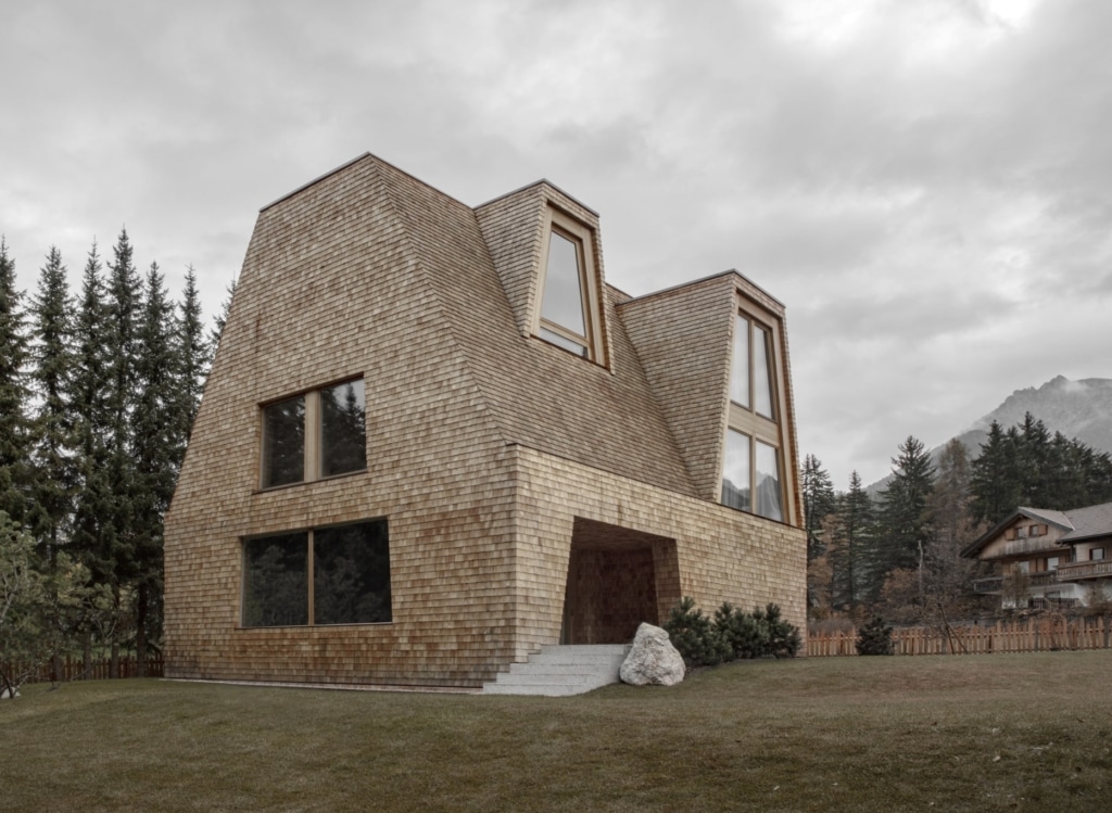 Una casa moderna con facciata in scandole di larice, situata in un paesaggio pittoresco. L’edificio presenta una forma geometrica distintiva con un tetto inclinato e grandi finestre che lasciano entrare molta luce naturale. In primo piano sono visibili una lastra di pietra e un grande masso che incorniciano l’ingresso della casa. Il giardino è curato e, sullo sfondo, si vedono alti abeti e altre abitazioni che creano un’atmosfera tranquilla e rurale. Il cielo è nuvoloso, il che sottolinea l’ambiente naturale.