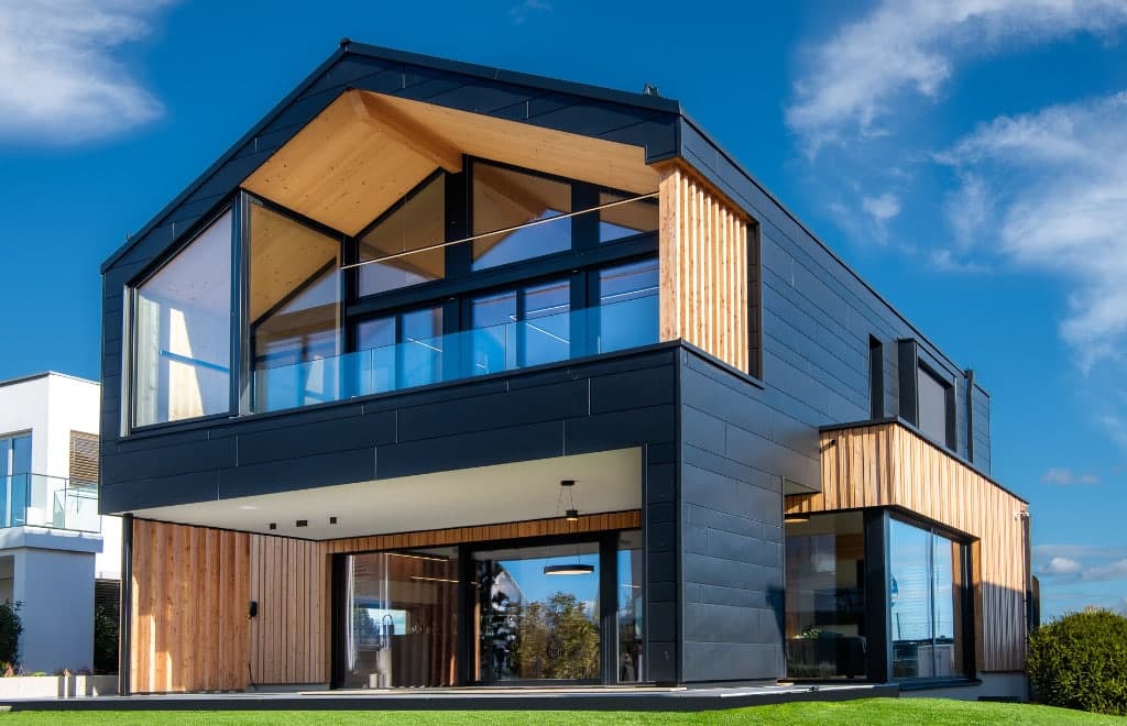 Una moderna casa modello realizzata con elementi in legno massello di holzius. L'edificio presenta una combinazione di rivestimenti in legno scuro e ampie vetrate che lasciano entrare molta luce naturale. La linea del tetto รจ inclinata e conferisce alla casa una silhouette distintiva. In primo piano si nota un prato ben curato, mentre l'architettura della casa si staglia chiaramente contro il cielo blu.