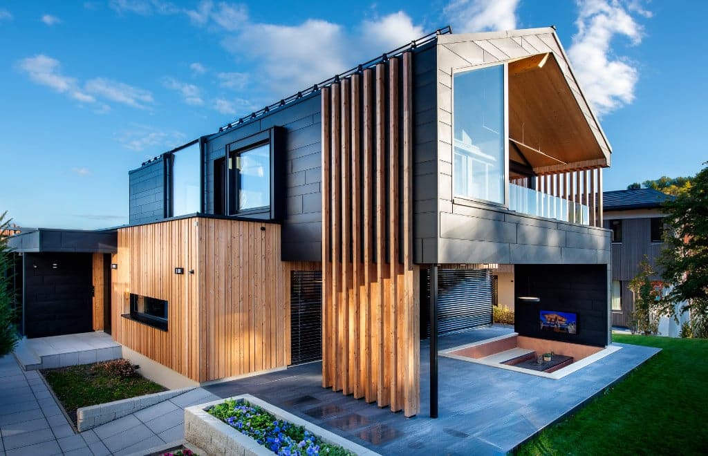 Una moderna casa modello di holzius, realizzata con pareti in legno massiccio. La facciata abbina un rivestimento in legno chiaro a elementi scuri e sobri e ampie finestre che lasciano entrare molta luce naturale. L'architettura รจ moderna e accattivante, con una linea del tetto inclinata e un balcone coperto. Davanti alla casa si estendono vialetti lastricati e un giardino ben curato con fiori colorati. Il cielo รจ blu con alcune nuvole bianche, il che crea un'atmosfera invitante.