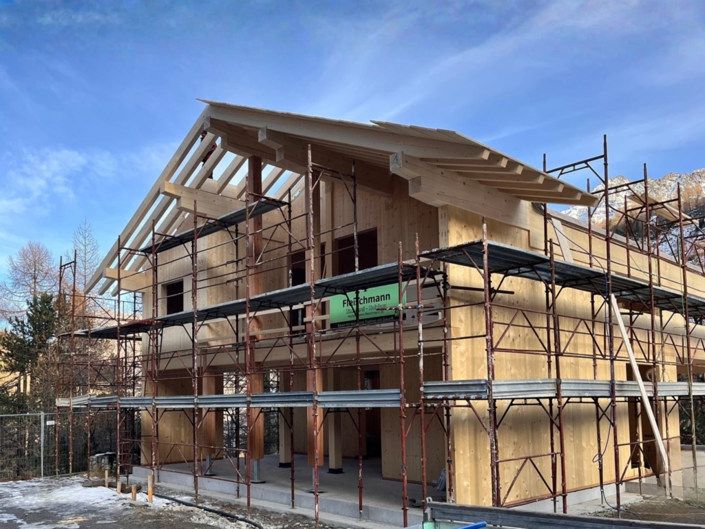 Casa in legno massiccio in fase di costruzione, circondata da ponteggi. Sono visibili le pareti in legno massiccio, gli elementi portanti del solaio e la struttura portante del tetto già montata.
