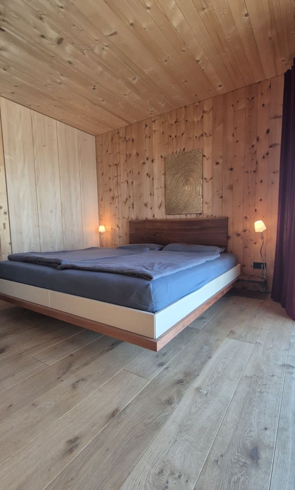 Una camera da letto luminosa con pavimento in legno e pareti e soffitto in legno dalle tonalità calde. Al centro della stanza si trova un letto sospeso con lenzuola blu, illuminato da due piccole lampade. Sopra il letto è appeso un quadro dorato. Sul lato sinistro si trova un armadio a muro con ante lisce in legno chiaro.