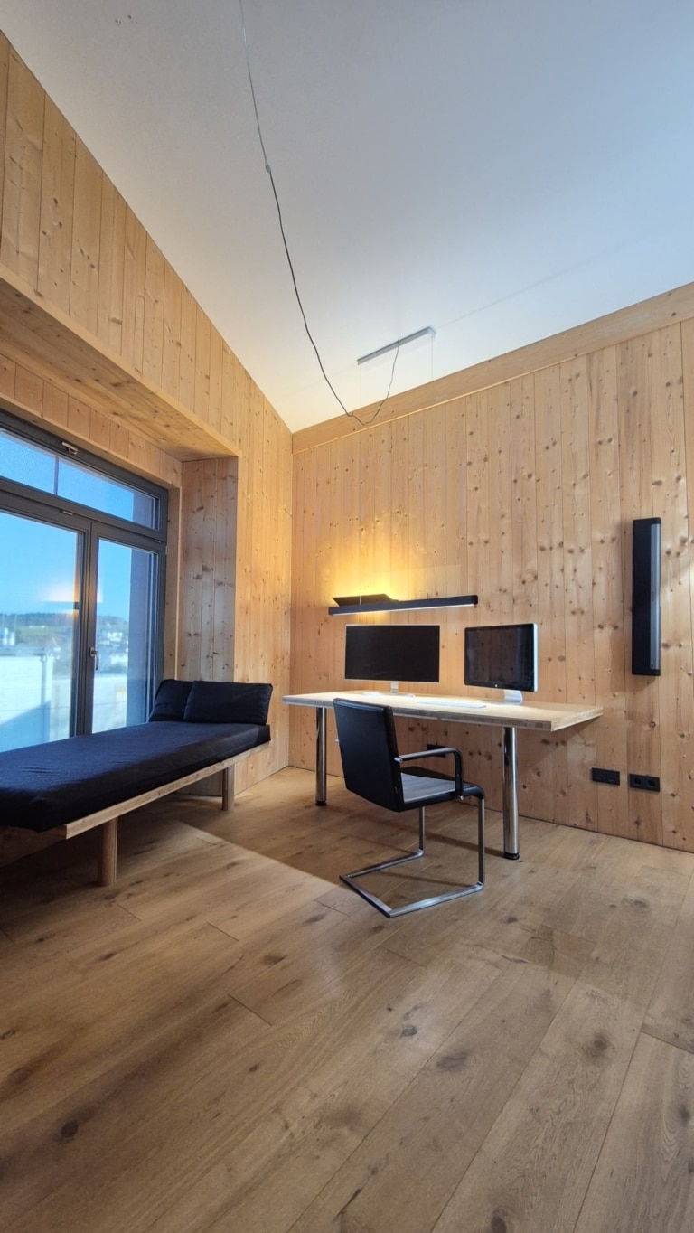 Interno di una stanza con pareti e soffitto in legno massello a vista di alta qualità. Si vedono una scrivania con monitor, una sedia, un divano vicino a una finestra e lampade moderne. Il design è contemporaneo e minimalista, con particolare enfasi sulla struttura naturale del legno.