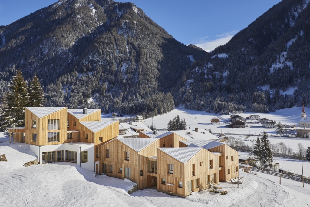 Un insieme di moderne case in legno con ripidi tetti a due falde, in parte coperti di neve, si adagia su un pendio verde davanti a uno scenario montano innevato sotto un cielo azzurro splendente. Le case sono rivestite con tavole di legno non trattato disposte verticalmente, che presentano venature naturali e un colore chiaro e caldo. La costruzione Holzius con gli elementi delle pareti è ben riconoscibile. La neve copre i tetti e il paesaggio circostante, conferendo alla scena un’atmosfera invernale e tranquilla. Una parte delle case è rifinita con intonaco bianco. Sullo sfondo si estende una valle innevata con altre case e un campanile. La combinazione di legno, neve e paesaggio montano conferisce all’insieme un carattere moderno e accogliente. La scena trasmette un senso di pace e di vicinanza alla natura. Alcuni alberi sono coperti di neve. Il sole splende intensamente creando giochi di luce e ombra sulle case e sulle montagne.