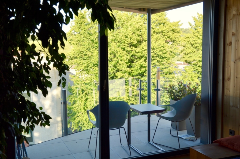 Una terrazza moderna con pareti in vetro e vista su un paesaggio verdeggiante. Sulla terrazza ci sono due sedie eleganti e un tavolino. Le piastrelle sono chiare e lisce, e lo spazio è circondato da piante che creano un'atmosfera accogliente. La luce del giorno filtra attraverso le ampie finestre, che offrono una vista sugli alberi all'esterno.
