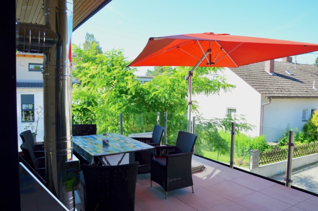 Una terrazza accogliente con un grande tavolo e diverse sedie, disposte sotto un ombrellone rosso brillante. Sullo sfondo si intravedono alberi verdi che incorniciano lo spazio esterno. La terrazza è pavimentata con piastrelle chiare e presenta una moderna ringhiera in vetro. Il tempo è soleggiato e sereno, il che crea un'atmosfera piacevole e invitante.