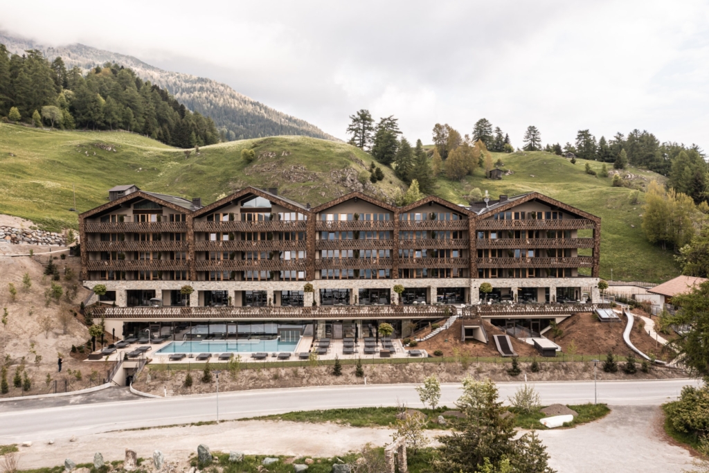 Vista esterna dell'hotel DAS GERSTL Family Retreat, immerso in un pittoresco paesaggio montano. La moderna facciata in legno e le ampie finestre conferiscono all'edificio un'atmosfera accogliente. In primo piano si nota un ampio giardino con sedie a sdraio e aree giochi per le famiglie. Il cielo sereno e i prati verdeggianti creano una rilassante atmosfera vacanziera.