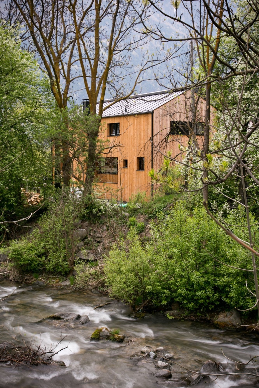 Una moderna casa in legno circondata da alberi rigogliosi e situata in riva a un piccolo fiume. La facciata della casa รจ costituita da pannelli di legno verticali, mentre le ampie finestre offrono una vista sulla natura circostante. Il fiume scorre dolcemente in primo piano, mentre il paesaggio รจ rigoglioso e verdeggiante, creando un'atmosfera tranquilla e naturale. Il cielo รจ nuvoloso, il che conferisce un'aria fresca.