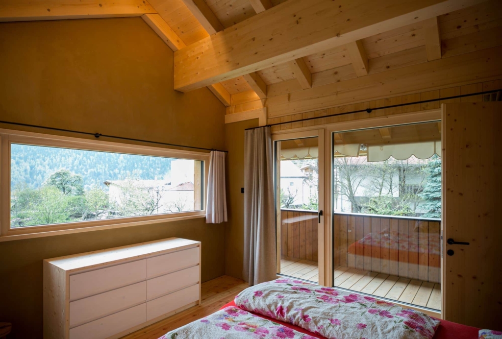 Una suggestiva immagine di una camera da letto con una parete in legno massello e un intonaco in argilla. In primo piano si vede un letto con un piumone colorato e dei cuscini. Sul lato destro c'รจ una grande porta a vetri che conduce a un balcone, mentre una finestra sul lato sinistro offre una vista sul paesaggio verdeggiante. Il soffitto รจ in legno con travi a vista, il che conferisce alla stanza un'atmosfera naturale e calda. Una cassettiera bianca รจ appoggiata alla parete.