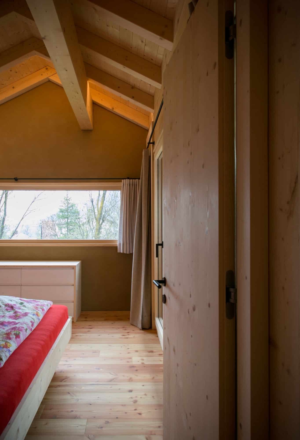 Una vista interna di una camera da letto con pareti intonacate in argilla dai caldi toni della terra. Il soffitto presenta travi a vista in legno che creano un'atmosfera accogliente. In primo piano si vede un letto con un copriletto rosso, mentre sul lato sinistro c'รจ una grande vetrata con tende che offre una vista sulla natura. Il pavimento รจ costituito da assi di legno chiaro che rendono l'ambiente ancora piรน arioso. Un armadio in legno รจ appoggiato alla parete, conferendo alla stanza un tocco naturale.