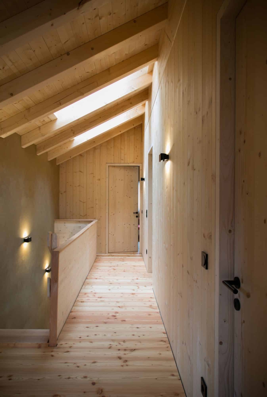 Un luminoso corridoio con pareti in legno massello e un soffitto in legno con travi a vista. Le pareti sono rivestite da pannelli di legno chiaro che mettono in risalto le venature naturali, mentre il pavimento รจ in parquet di legno massello chiaro. Sulla parete di sinistra si notano due porte, mentre sul lato destro una ringhiera in legno costeggia la zona delle scale. Due piccole applique illuminano delicatamente il corridoio. Una finestra sul tetto lascia inoltre entrare la luce del giorno nella stanza.
