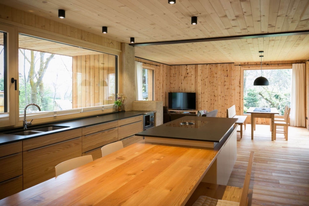Una cucina moderna con pareti e soffitto in legno di cembro chiaro. Le pareti in legno massello presentano una venatura naturale, mentre il soffitto in legno รจ dotato di luci a LED integrate. In primo piano spicca una grande isola da cucina in legno con piano cottura nero. Ampie finestre lasciano entrare la luce del giorno e offrono una vista sul verde circostante. Sullo sfondo si intravedono un tavolo da pranzo con sedie e un televisore, che creano un'atmosfera accogliente e invitante.