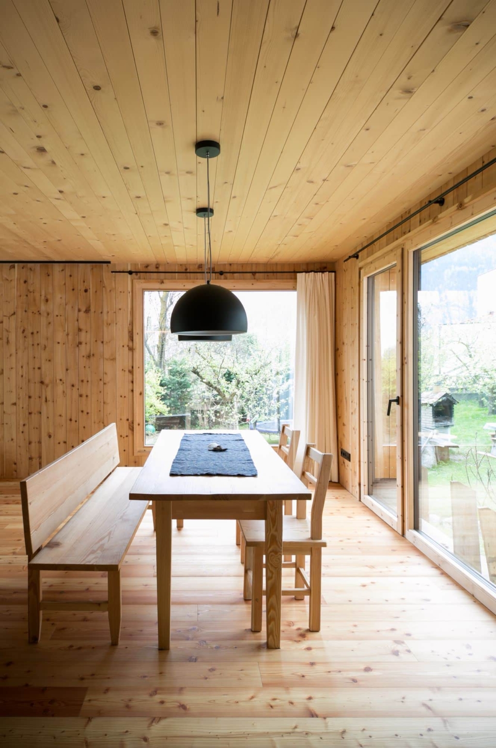 Una cucina moderna con un soffitto in legno massello composto da pannelli chiari, che crea un'atmosfera calda e accogliente. La cucina dispone di un'elegante isola con piano cottura nero e piano di lavoro in legno. Sulla parete sono presenti ampie finestre che lasciano entrare molta luce naturale.
