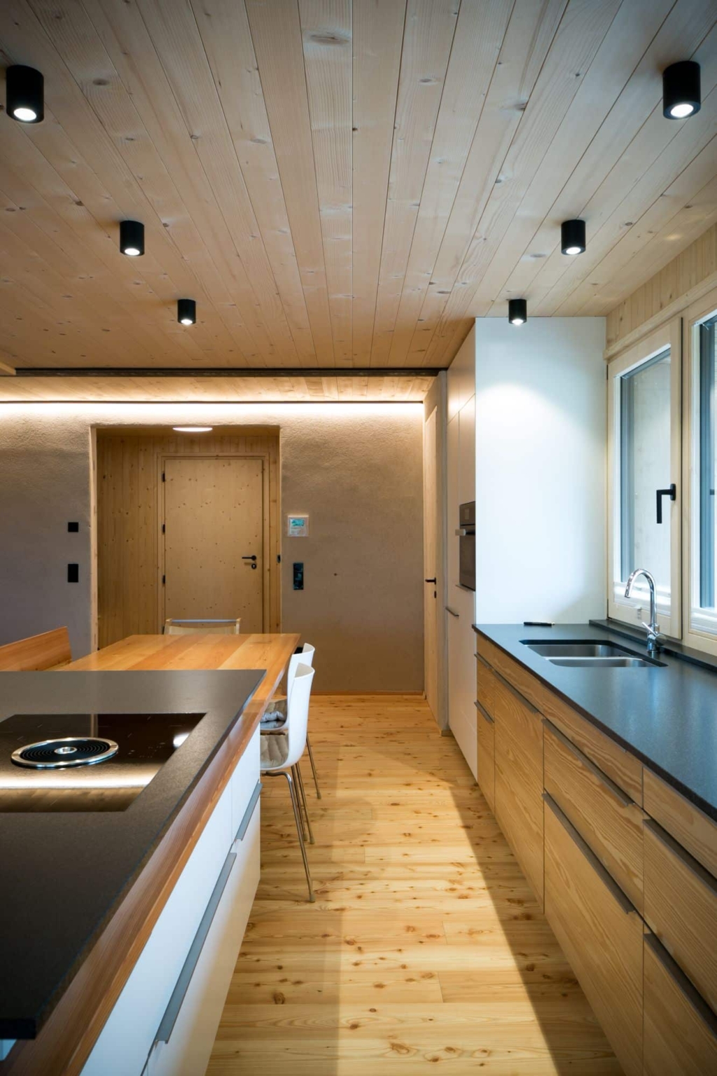 Una cucina moderna con un soffitto in legno massello composto da pannelli chiari, che crea un'atmosfera calda e accogliente. La cucina dispone di un'elegante isola con piano cottura nero e piano di lavoro in legno. Sulla parete sono presenti ampie finestre che lasciano entrare molta luce naturale. I mobili sono in legno chiaro e l'illuminazione รจ costituita da moderni faretti da incasso che illuminano delicatamente l'ambiente. Anche il pavimento รจ in legno chiaro, il che rafforza l'armoniosa impressione d'insieme.