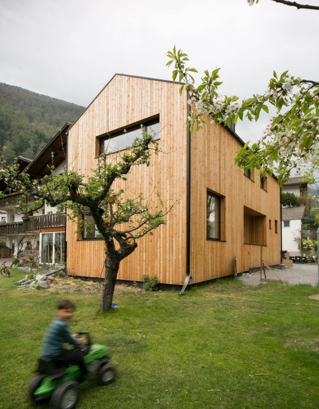 Una moderna casa in legno massiccio realizzata da holzius, con una facciata composta da listelli di legno disposti verticalmente. L'edificio presenta un tetto spiovente e ampie finestre che lasciano entrare molta luce naturale. In primo piano si vede un giardino ben curato con un piccolo albero in fiore e un prato su cui un bambino guida un veicolo giocattolo verde. L'ambiente รจ rurale, con dolci colline sullo sfondo, e trasmette un'atmosfera accogliente.