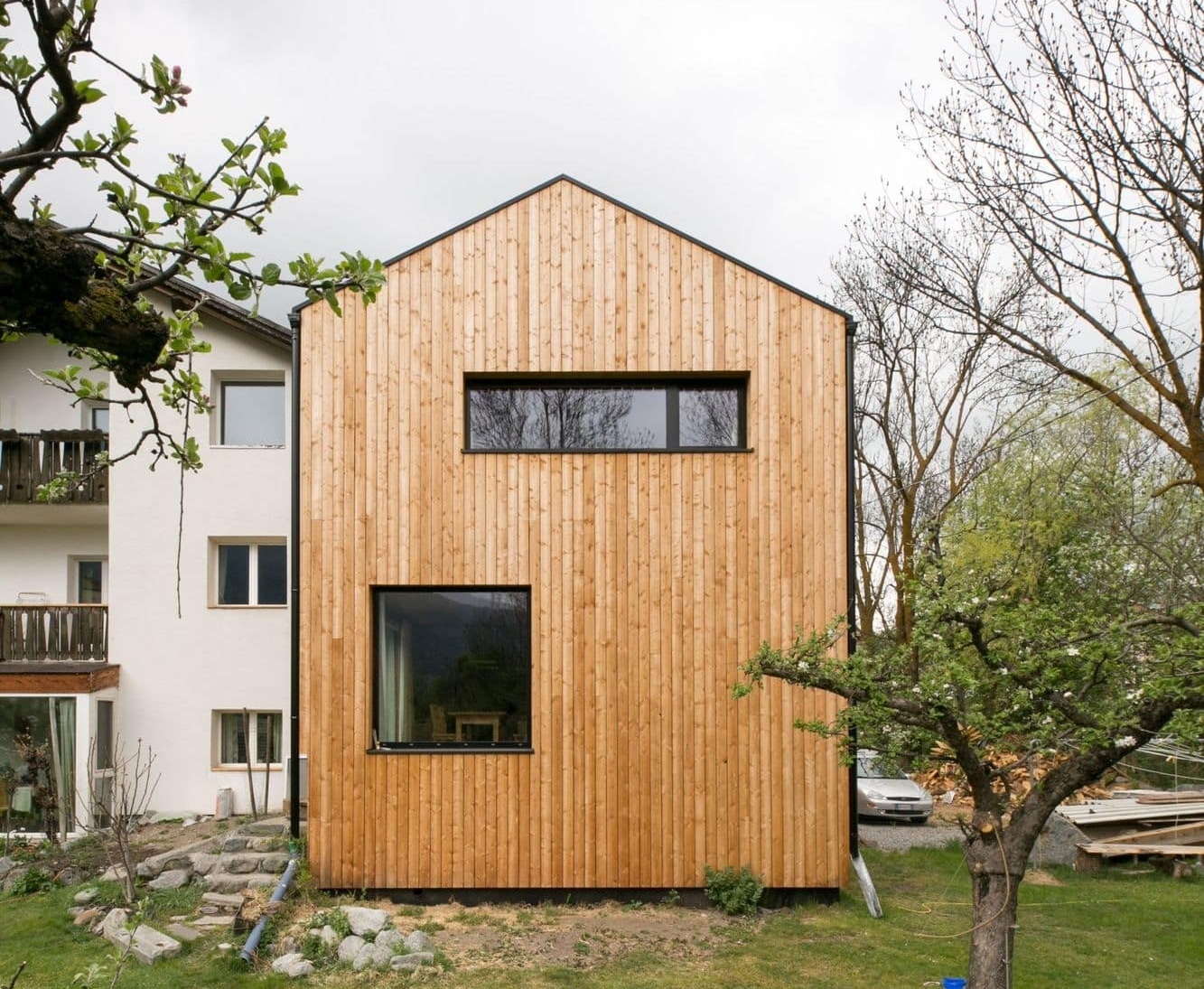 Una moderna casa in legno massiccio Holzius con una facciata composta da listelli di legno disposti verticalmente. L'edificio presenta un tetto spiovente e ampie finestre che lasciano entrare molta luce naturale. In primo piano si intravede un piccolo giardino con alcuni alberi e un vialetto lastricato. Sul lato sinistro si trova un altro edificio che crea un contrasto con il rivestimento in legno della nuova casa. Il cielo รจ nuvoloso, il che crea un'atmosfera tranquilla e naturale.