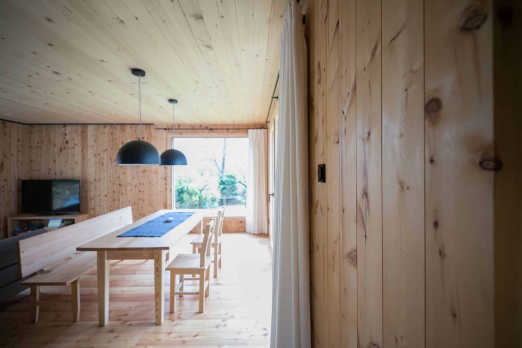 Una vista interna di una zona pranzo con pareti in legno di cembro chiaro. Le pareti in legno massello presentano venature naturali e nodi. In primo piano si trova un lungo tavolo in legno con una tovaglia blu, circondato da sedie chiare. Sopra il tavolo sono appese due lampade a sospensione nere. In un angolo si intravede un televisore, mentre ampie finestre lasciano entrare la luce del giorno, conferendo alla stanza un'atmosfera accogliente e invitante.