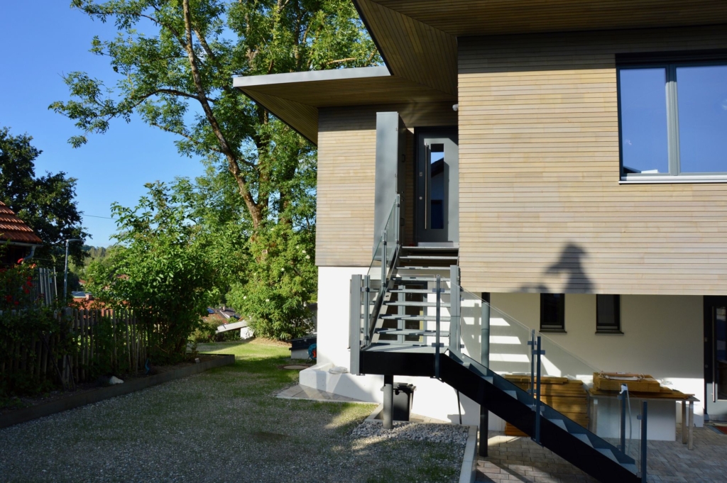 Una vista esterna di una casa moderna con facciata in legno e tetto spiovente. Una scala in metallo conduce alla porta d'ingresso, situata su una veranda rialzata. Il giardino è ben curato e pavimentato con ghiaia ed erba. Sullo sfondo si intravedono alberi verdi che creano un'atmosfera naturale. Il cielo è limpido e azzurro, il che sottolinea l'atmosfera accogliente della casa.