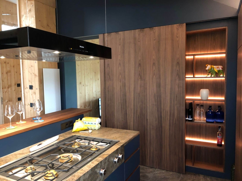 Una cucina moderna caratterizzata da una combinazione di superfici in legno e blu. In primo piano si trova un piano cottura sormontato da una cappa in acciaio inossidabile. Sul lato sinistro, alcuni bicchieri sono disposti su un ripiano in legno. La parete dietro il piano cottura è rivestita in legno, mentre una mensola a muro con illuminazione integrata sul lato destro espone diverse bottiglie e oggetti decorativi. L'atmosfera è elegante e accogliente, con un mix armonioso di materiali.