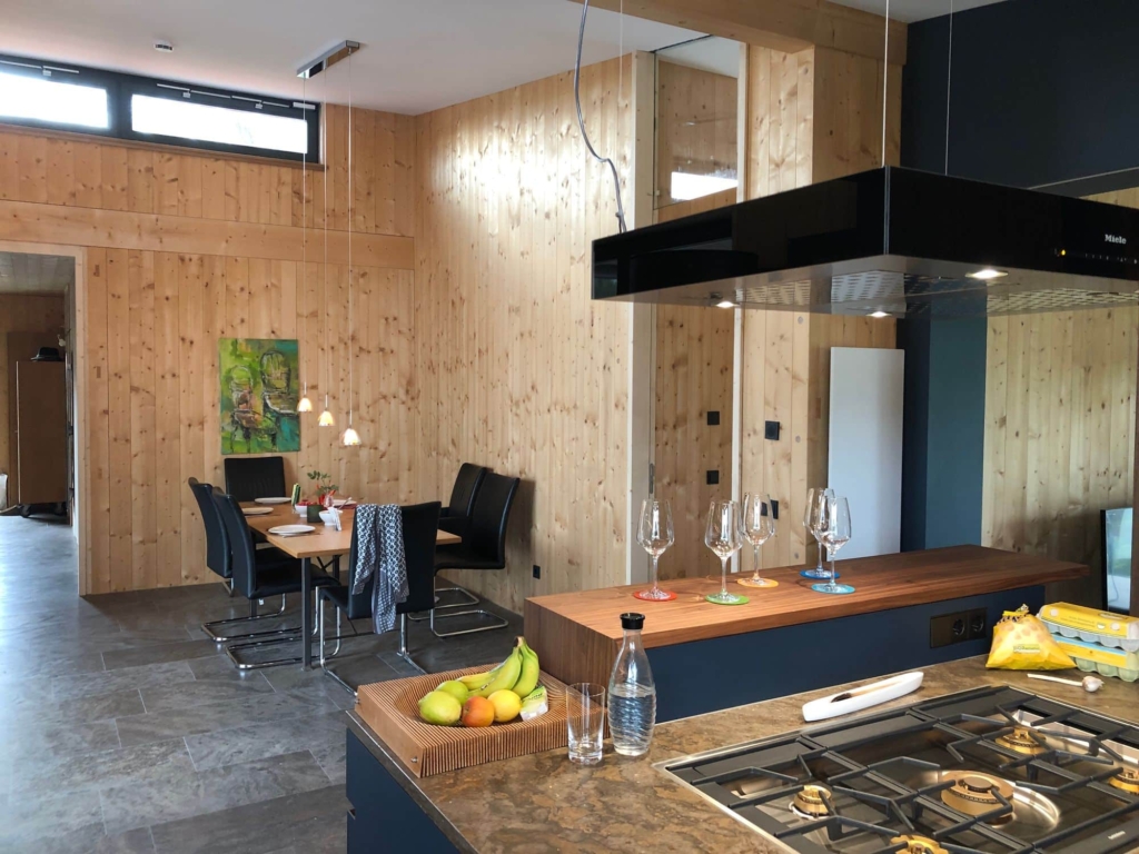Una vista interna di un soggiorno moderno con pareti rivestite di pannelli in legno chiaro. Sul lato sinistro si vede un tavolo da pranzo con sedie nere, decorato con un vaso di fiori. In primo piano spicca un'isola da cucina con piano di lavoro in granito e piano cottura integrato. Sopra l'isola da cucina sono appese moderne lampade a sospensione. Le ampie finestre lasciano entrare la luce del giorno e creano un'atmosfera accogliente. Il pavimento è in piastrelle grigie, che creano un piacevole contrasto con le superfici in legno.