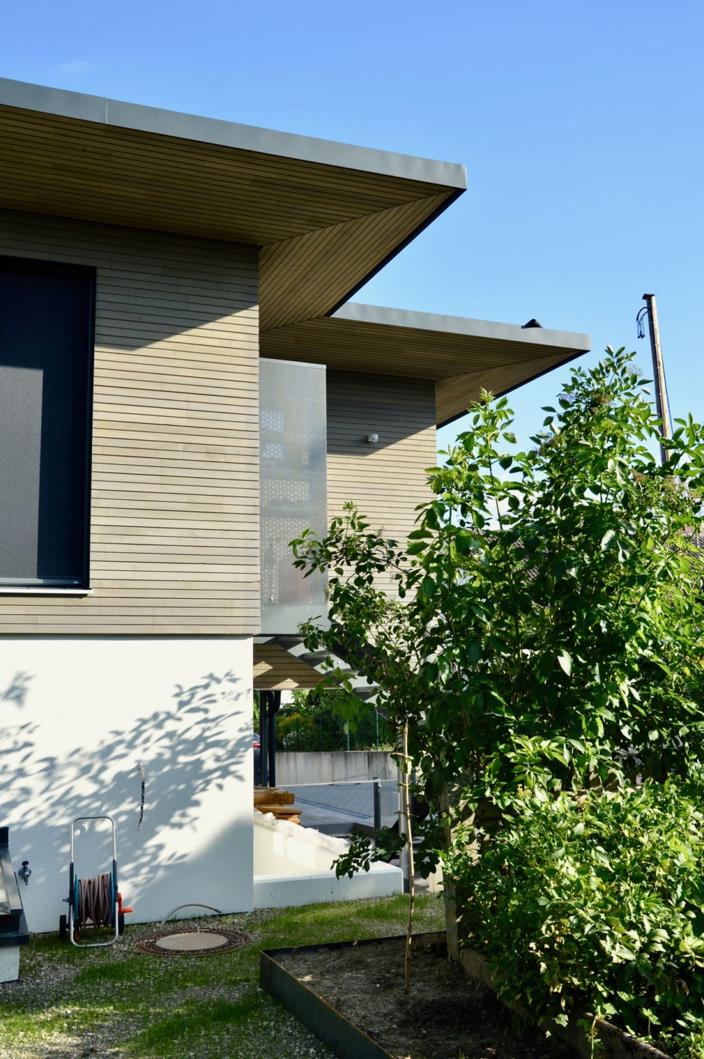 Una vista esterna di una casa moderna con rivestimento in legno e tetto spiovente. La facciata presenta ampie finestre e una terrazza coperta. In primo piano si vedono piante verdi e arbusti che creano un ambiente naturale. La luce del sole illumina delicatamente la struttura, mentre il cielo azzurro e limpido sullo sfondo crea un'atmosfera accogliente.