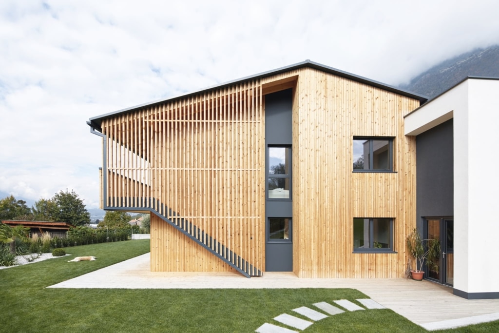 Una moderna casa in legno massiccio Holzius con una facciata composta da assi di legno chiare disposte verticalmente e un tetto a due falde. Una scala esterna con ringhiera grigia conduce a un balcone rivestito con doghe di legno. Le finestre sono di colore grigio. La tecnica costruttiva con elementi per pareti e soffitti Holzius è qui ben riconoscibile. Sullo sfondo si stagliano le montagne. Un prato ben curato si estende intorno alla casa. Un moderno annesso con intonaco bianco e dettagli grigi completa l'insieme.