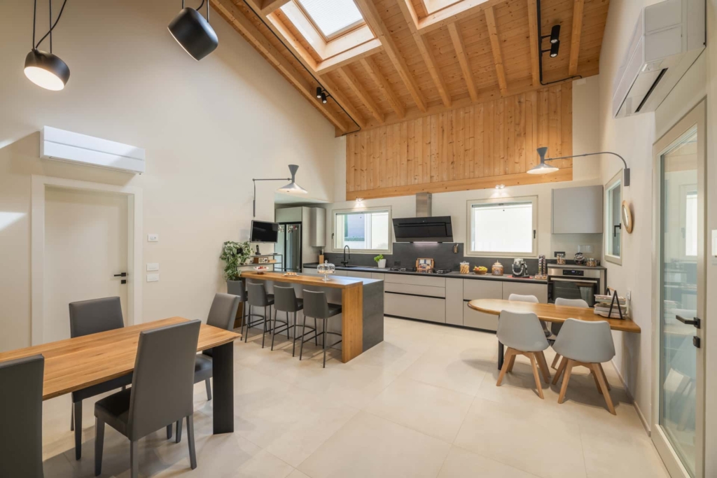 Una zona soggiorno-cucina luminosa e spaziosa con un alto soffitto a volta in legno massello Holzius, interrotto da diversi lucernari. Lo stile costruttivo Holzius, con i suoi elementi per soffitti, รจ ben riconoscibile. La stanza presenta un pavimento in legno e pareti bianche. Nella zona pranzo si trova un tavolo in legno con sedie grigie. In primo piano si trova un'isola da cucina con piano in legno e sgabelli da bar grigi. Sullo sfondo si trova una cucina moderna con mobili grigi. Lampade a sospensione illuminano la stanza. Una TV a schermo piatto รจ appesa alla parete. La combinazione di legno ed elementi moderni conferisce alla stanza un tocco elegante e accogliente. Un condizionatore รจ montato a parete.