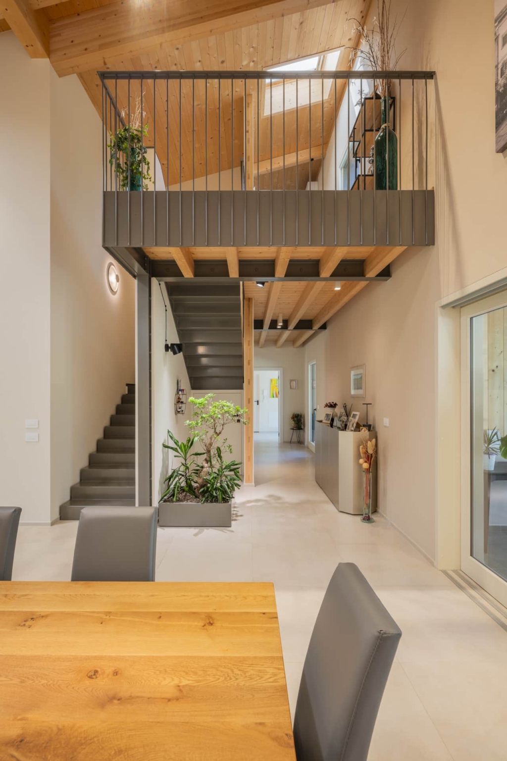 Una zona giorno luminosa e aperta con un soffitto in legno massello Holzius che si estende fino al frontone ed รจ interrotto da un lucernario. Una scala grigia conduce a una galleria con una ringhiera in metallo nero. In primo piano si trova un grande tavolo in legno. Sedie grigie circondano il tavolo. Una pianta da appartamento in un vaso grigio si trova accanto alla scala. La luce del giorno entra attraverso una porta in fondo alla stanza. La tecnica costruttiva Holzius con pareti in legno massiccio e soffitto in legno massiccio crea un'atmosfera calda e accogliente. Gli elementi delle pareti Holzius sono parzialmente visibili.