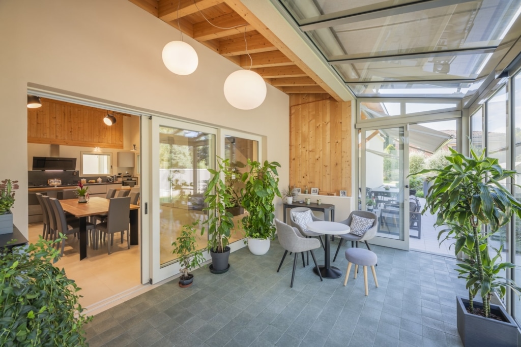 Un giardino d'inverno con tetto e pareti in vetro, rivestito con piastrelle nei toni del grigio. Una porta scorrevole aperta conduce a una stanza adiacente con pareti in legno e un tavolo da pranzo con sedie. Diverse piante da appartamento in vaso sono disposte nel giardino d'inverno e conferiscono all'ambiente un tocco naturale. Nella parte posteriore del giardino d'inverno si trova un piccolo tavolo rotondo con sedie e uno sgabello. Due lampade sferiche pendono dal soffitto. Lo stile architettonico Holzius potrebbe riflettersi nella struttura in legno della stanza adiacente. La luce del giorno inonda la stanza creando un'atmosfera luminosa e accogliente.