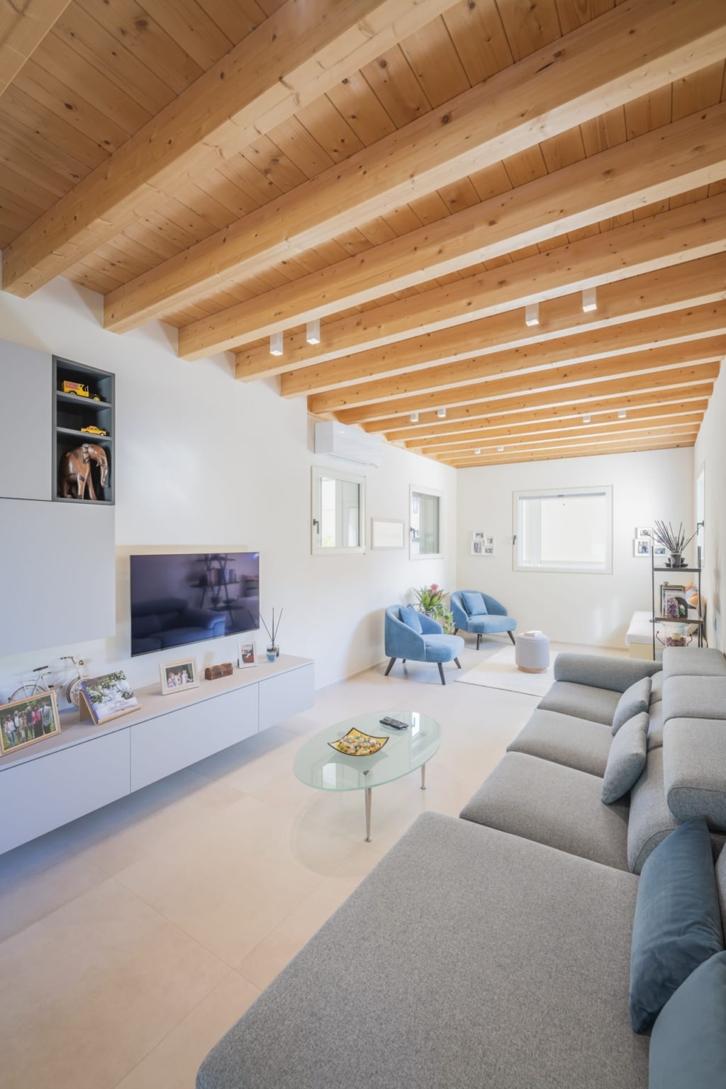 Un soggiorno luminoso e moderno con soffitto in legno massello Holzius, composto da travi di legno chiaro. Le pareti sono intonacate di bianco. Un divano ad angolo grigio domina la stanza. Davanti al divano si trova un tavolino ovale in vetro. Contro la parete รจ posizionato un mobile basso con un televisore. Sullo sfondo si trovano due poltrone azzurro chiaro. Lo stile Holzius con le travi in legno crea un'atmosfera calda e accogliente. La combinazione di legno ed elementi moderni conferisce alla stanza un tocco elegante e confortevole.