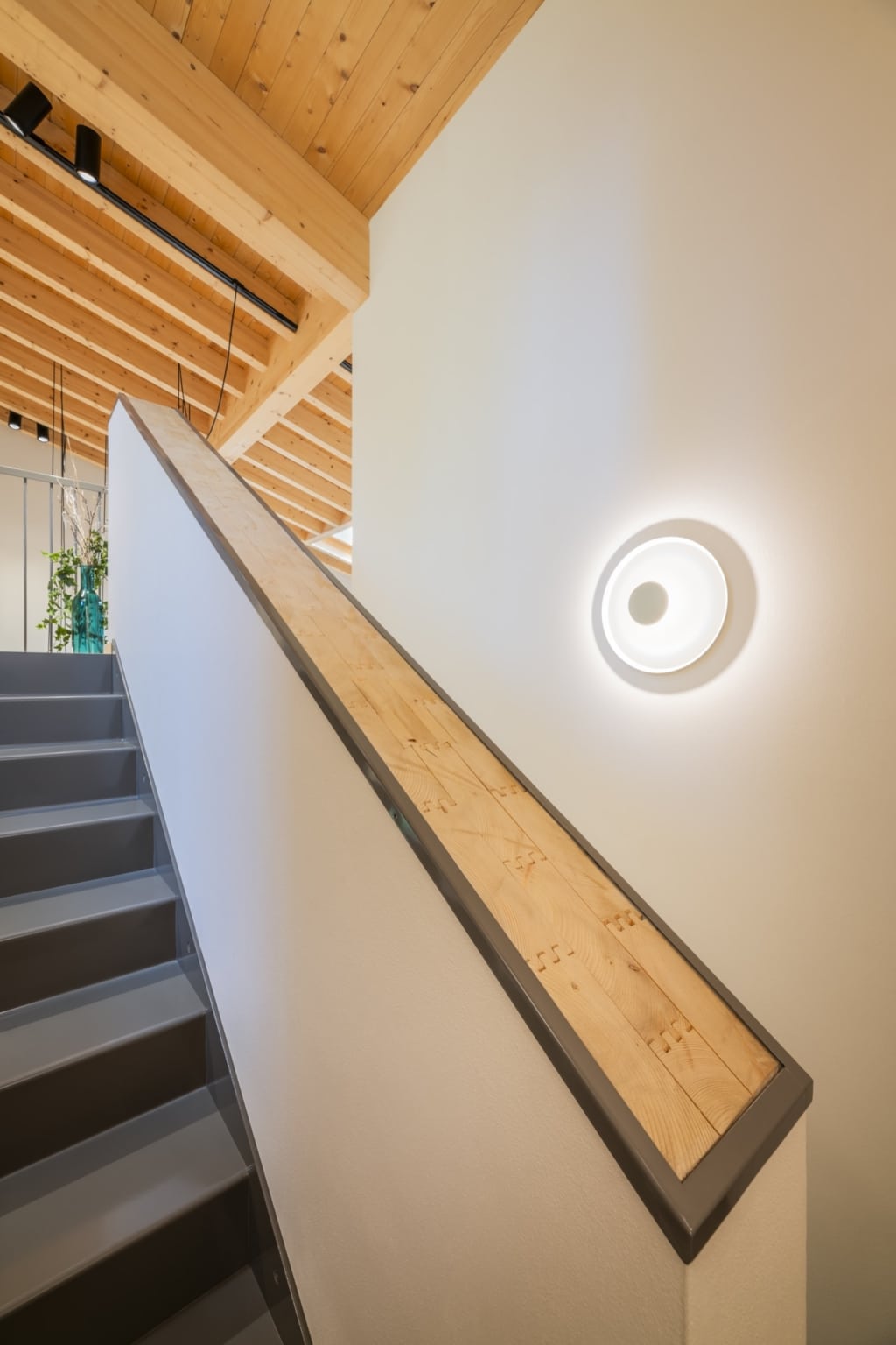 Una scala in una casa in legno massiccio Holzius con un soffitto in legno massiccio Holzius rivestito con pannelli di legno chiaro non trattato. La scala stessa รจ verniciata di grigio e presenta una ringhiera con un corrimano in legno incorniciato da una struttura metallica grigia. Una lampada da parete rotonda e moderna proietta una luce calda sulla parete bianca. La tecnica costruttiva Holzius, con pareti in legno massiccio e soffitto in legno massiccio, crea un'atmosfera calda e accogliente. La combinazione di legno, metallo e linee pulite conferisce alla stanza un tocco moderno e naturale. Sullo sfondo si intravede una parte di una stanza con piante d'appartamento. Gli elementi delle pareti Holzius sono parzialmente visibili.