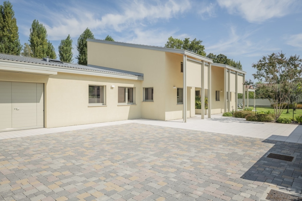 Una casa moderna di colore beige con tetto piano e un piazzale pavimentato. La casa dispone di un garage con porta avvolgibile e diverse finestre rettangolari. L'ingresso รจ sostenuto da colonne. Il giardino รจ abbellito da alberi, cespugli e un prato ben curato. Il cielo รจ blu con qualche nuvola sparsa. Lo stile architettonico Holzius potrebbe riflettersi nella struttura del tetto e delle colonne. La combinazione di intonaco, legno e vetro conferisce alla casa un tocco moderno ed elegante. Il piazzale pavimentato รจ realizzato con pietre di diversi colori.