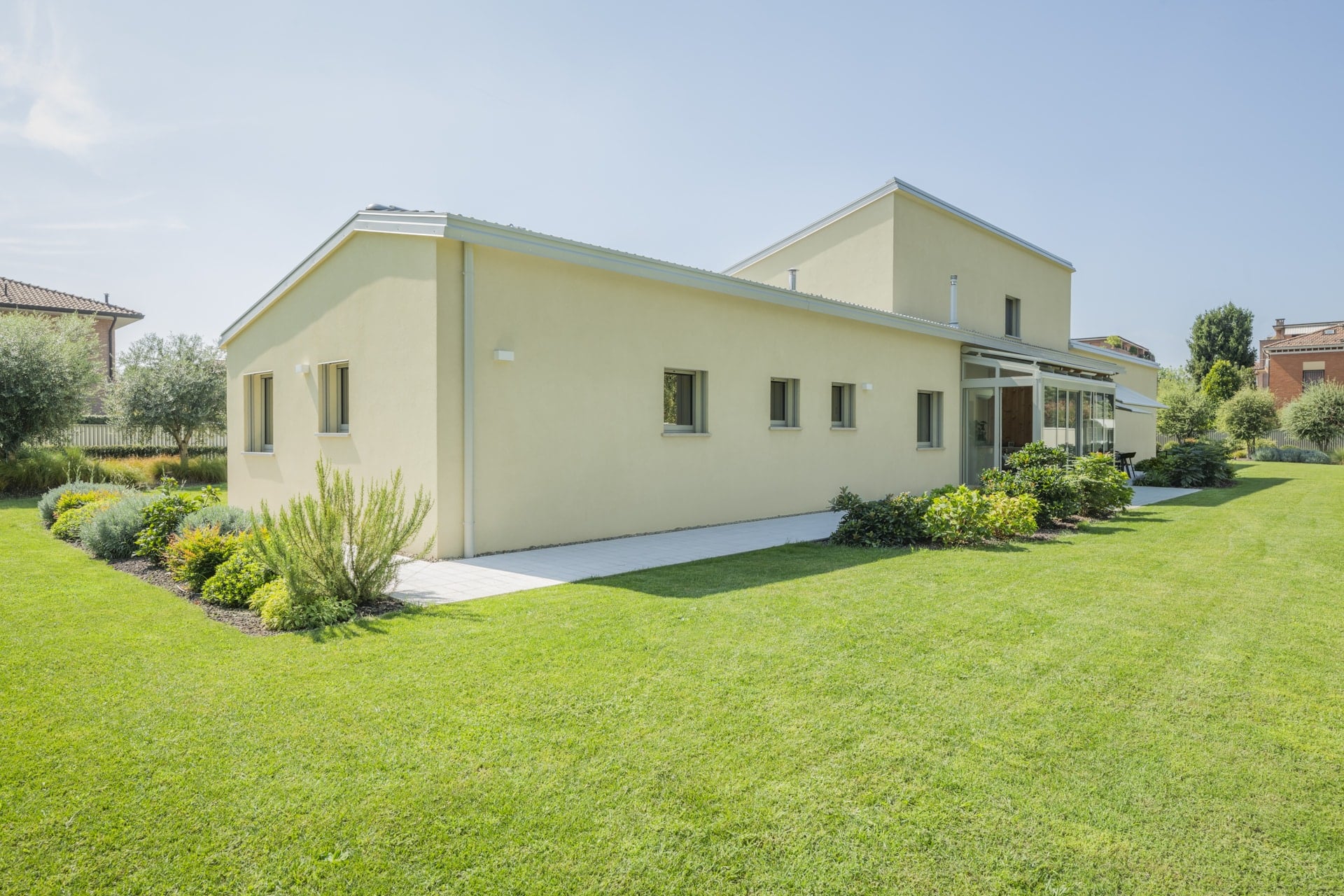 Una casa moderna a un piano con una facciata giallo chiaro e un tetto piano, circondata da un prato verde e da una vegetazione rigogliosa. Le finestre sono rettangolari e hanno infissi scuri. Una veranda vetrata con porte scorrevoli in vetro collega le due parti della casa. Un vialetto stretto e bianco costeggia la facciata. Lo stile architettonico Holzius potrebbe riflettersi nella costruzione della casa, in particolare nell'uso del legno per gli elementi portanti. Il giardino รจ abbellito da varie piante, cespugli e alberi. La scena trasmette un'atmosfera tranquilla e a contatto con la natura. Sullo sfondo si intravede un'altra casa con un tetto di tegole rosse.