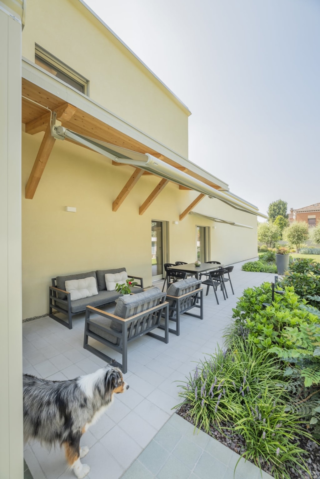 Una casa a due piani con una facciata giallo chiaro e una terrazza coperta. Lo stile Holzius potrebbe riflettersi nella struttura della copertura. Una tenda da sole รจ fissata alla facciata e offre ombra. Sulla terrazza si trovano diversi mobili da giardino grigi con cuscini grigi: un divano, una poltrona e un tavolo. Sullo sfondo c'รจ un tavolo da pranzo con sedie. In primo piano c'รจ un cane dal pelo grigio e marrone. Il pavimento della terrazza รจ rivestito con piastrelle grigio chiaro. Una rigogliosa vegetazione con cespugli verdi e fiori viola si trova sulla destra dell'immagine. Gli elementi di rivestimento Holzius potrebbero essere nascosti dietro la facciata intonacata. La combinazione di legno, intonaco e mobili moderni conferisce alla casa un tocco elegante e accogliente.