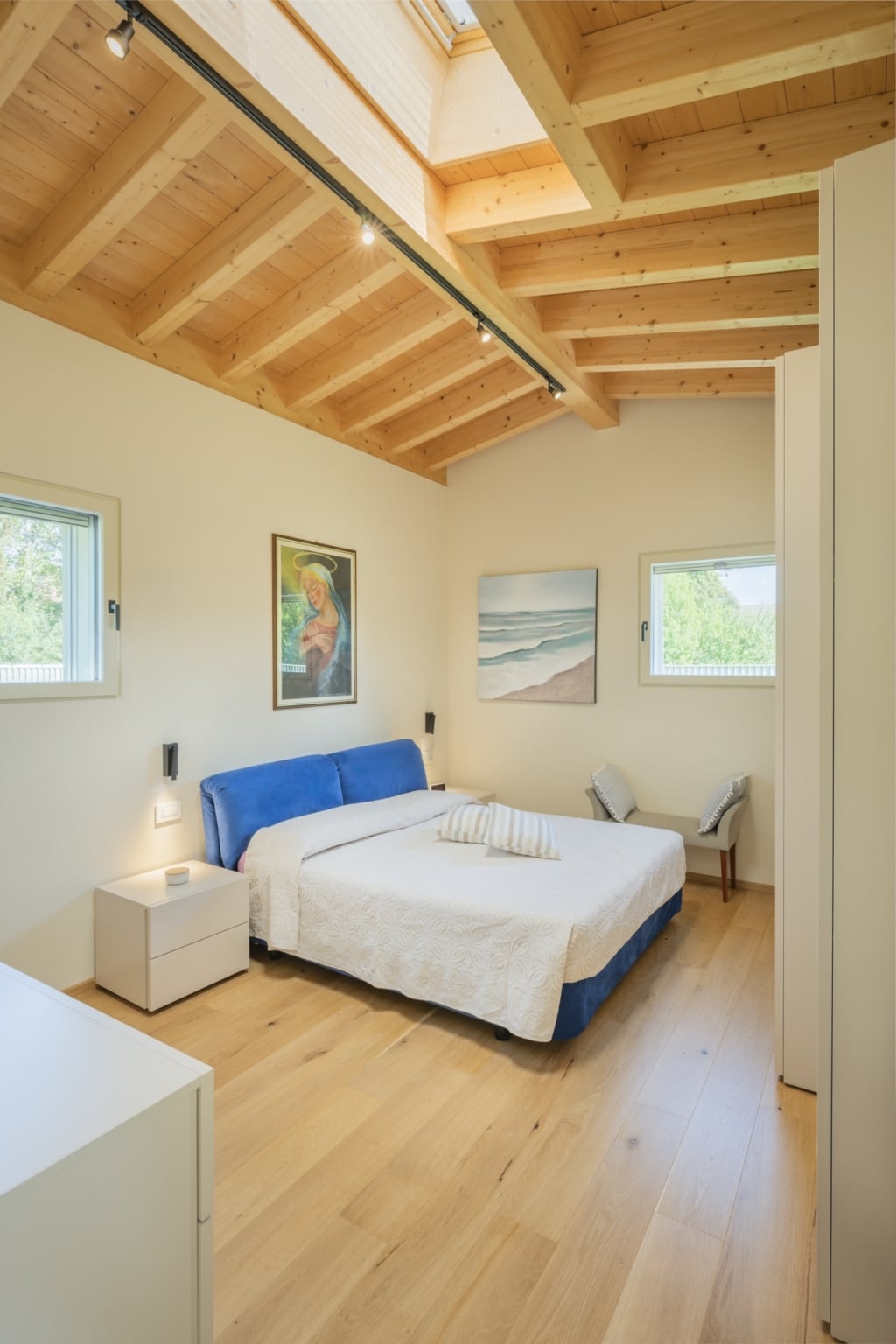 Una camera da letto luminosa e moderna con un soffitto in legno massello Holzius composto da travi chiare e un pavimento in legno. Le pareti sono dipinte in una delicata tonalitร crema. Nella stanza si trova un grande letto blu con un copriletto bianco e un cuscino allungato. Due piccoli comodini bianchi sono posizionati accanto al letto. Sopra i comodini sono appese delle moderne lampade da parete. Alle pareti sono appesi due quadri incorniciati. Una finestra stretta con telaio in legno si trova su entrambi i lati del letto. Un lucernario nel soffitto lascia entrare molta luce naturale nella stanza. La struttura Holzius con pareti in legno massello e soffitto in legno massello crea un'atmosfera calda e accogliente. Sullo sfondo si trova una sedia con un cuscino grigio. La combinazione di legno ed elementi moderni conferisce alla stanza un tocco elegante e accogliente.