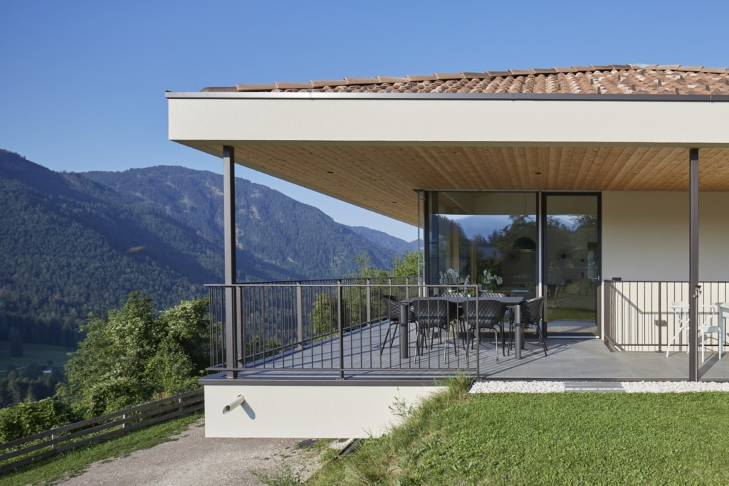 Una casa moderna con tetto in tegole e un balcone coperto con elementi per soffitti Holzius. Il balcone è circondato da una ringhiera grigia e offre una vista panoramica su un paesaggio verdeggiante con montagne boscose. Sul balcone sono presenti un tavolo e delle sedie. Una grande porta scorrevole in vetro conduce all'interno della casa. Lo stile costruttivo Holzius è riconoscibile dagli elementi per soffitti Holzius. La casa si integra armoniosamente nell'ambiente naturale.