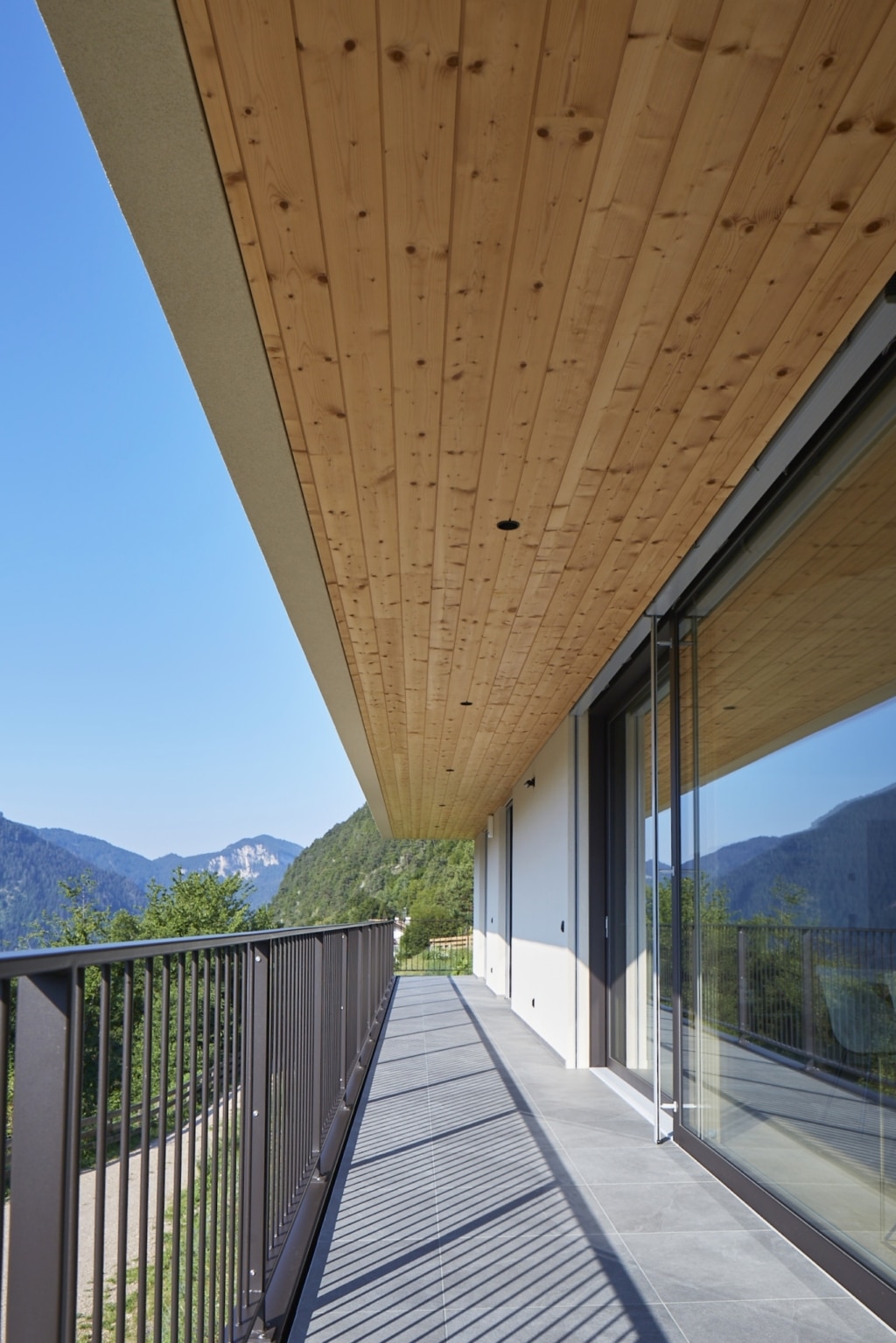 Un balcone lungo e stretto con elementi a soffitto Holzius e pavimento in piastrelle grigie. Una ringhiera grigia e sottile con montanti verticali corre lungo il perimetro esterno del balcone. Una grande porta scorrevole in vetro conduce all’interno della casa e riflette il paesaggio verdeggiante. Sullo sfondo si ergono colline boscose sotto un cielo blu. Lo stile architettonico Holzius è riconoscibile dall'uso del legno. La combinazione di legno, vetro e metallo conferisce al balcone un tocco moderno e accogliente. Le ombre della ringhiera creano un motivo interessante sul pavimento.