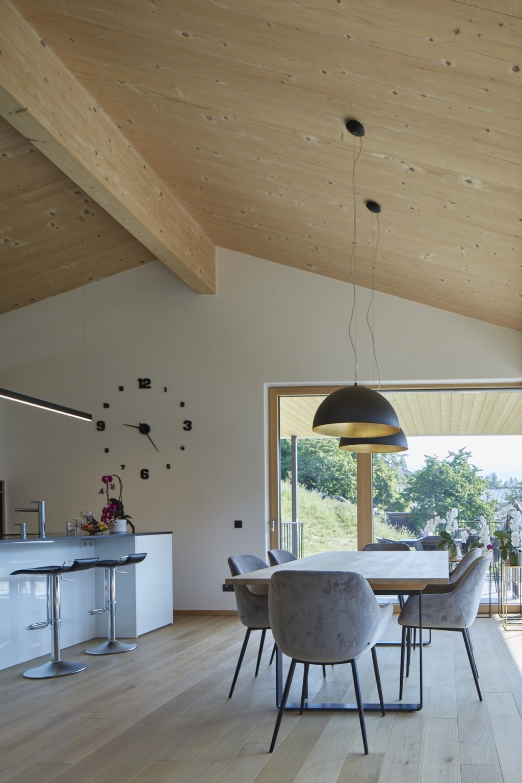 Una zona pranzo luminosa e moderna con pavimento in legno e un soffitto in legno massello Holzius composto da pannelli chiari che si estendono fino al frontone. Lo stile Holzius si riconosce dall’uso del legno. Al centro della stanza si trova un tavolo in legno con sedie grigie. Due moderne lampade a sospensione con l’esterno nero e l’interno dorato illuminano il tavolo. Un grande orologio con numeri neri è appeso alla parete. Un'isola da cucina con ante bianche e due sgabelli da bar si trova sulla sinistra dell'immagine. Una vetrata con una grande finestra offre una vista panoramica sul paesaggio verde. La combinazione di legno ed elementi moderni conferisce alla stanza un tocco elegante e accogliente.
