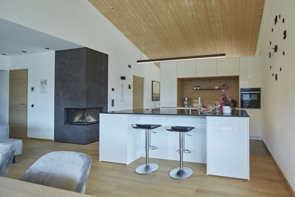 Una zona soggiorno-cucina luminosa e moderna con un soffitto in legno massello Holzius rivestito con pannelli in legno chiaro. Il pavimento è rivestito con assi di legno chiaro. Un camino grigio domina la stanza e funge da divisorio. In primo piano spicca un'isola da cucina con piano di lavoro nero e ante bianche. Due sgabelli da bar in metallo sono posizionati accanto all'isola. Sullo sfondo si trova una cucina a vista con mobili bianchi e una nicchia con elementi in legno. Un moderno elemento di illuminazione a forma di linea orizzontale pende dal soffitto. Un grande orologio con cifre e lancette nere è appeso alla parete. Lo stile Holzius crea un'atmosfera calda e accogliente. La combinazione di legno ed elementi moderni conferisce alla stanza un tocco elegante e funzionale.